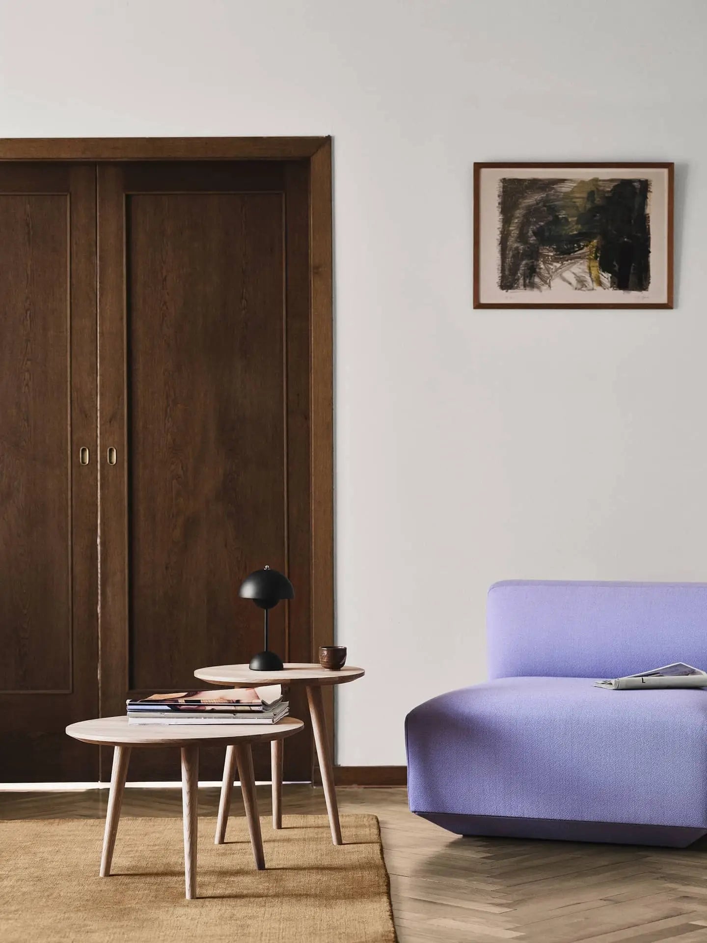 A modern living room with a light purple sofa, two round wooden coffee tables topped with books and an &Tradition Flowerpot VP9 Portable Table Lamp, a brown rug, wooden sliding doors, and an abstract painting on a white wall.