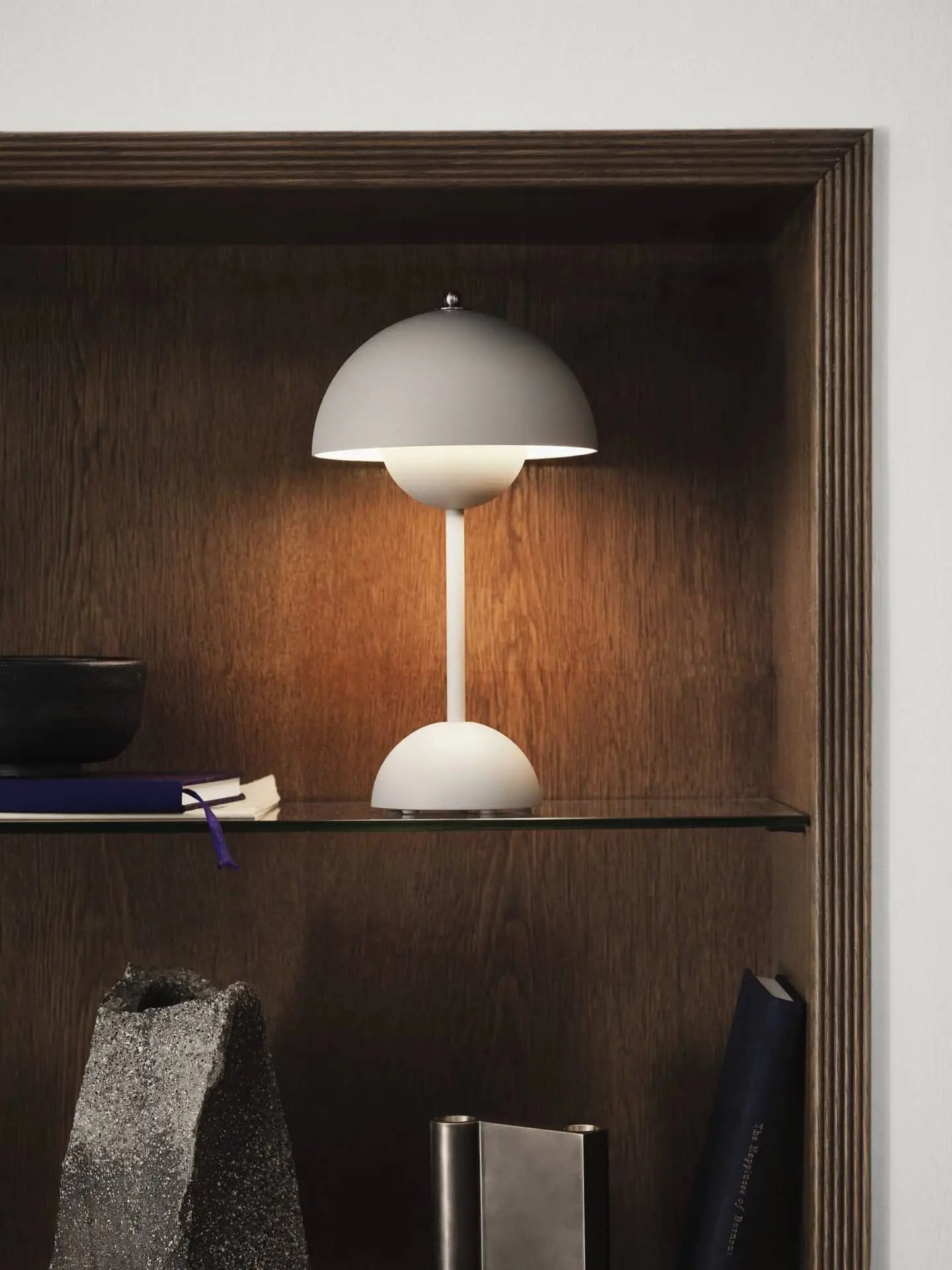 The &Tradition Flowerpot VP9 Portable Table Lamp, with its modern white dome-shaped shade, sits on a glass shelf in a wooden alcove among books, a bowl, and a stone sculpture. Its turned on and comes with a USB charging cable.