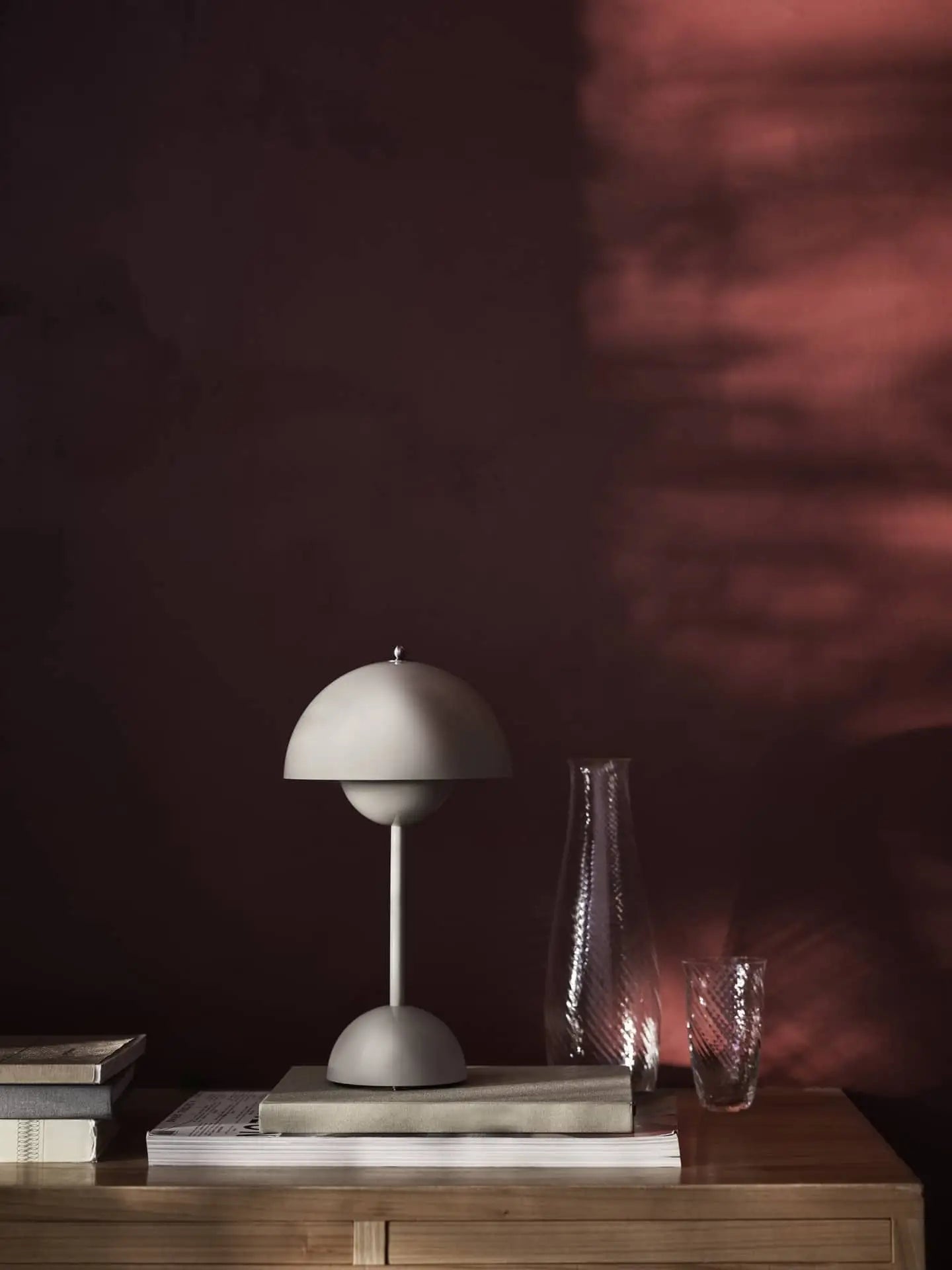 A minimalist scene featuring the &Tradition Flowerpot VP9 Portable Table Lamp, a glass carafe, and tumbler on wood with stacked books beside them. Warm light from the lamp casts soft shadows on a dark maroon background.