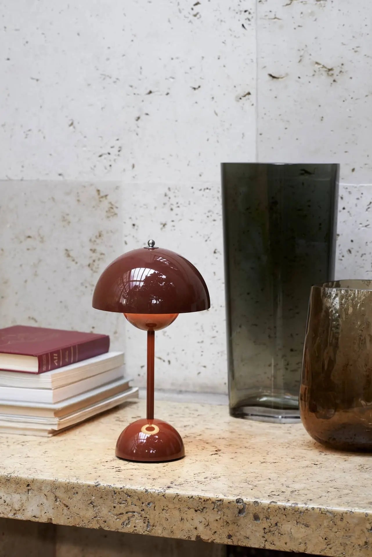 The &Tradition Flowerpot VP9 Portable Table Lamp, a small brown dome-shaped piece, sits on marble beside books and two tall glass vases—one clear grey, one brown—against a textured white stone wall. Includes USB charging cable.