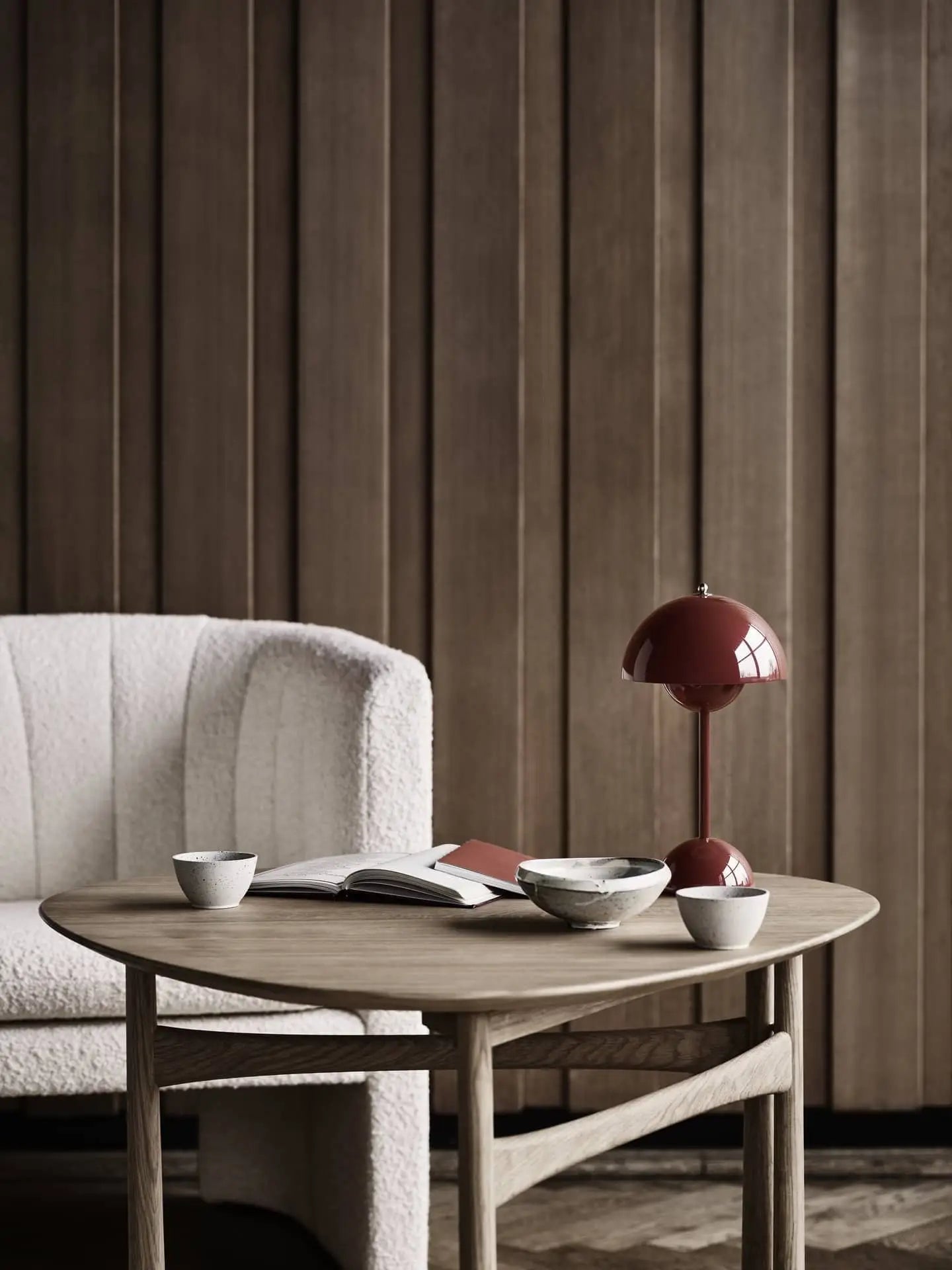 A minimalist living space with a light, textured armchair, a wooden table holding ceramic cups and an open book, and an &Tradition Flowerpot VP9 Portable Table Lamp with USB charging cable, set against vertical wooden panels.