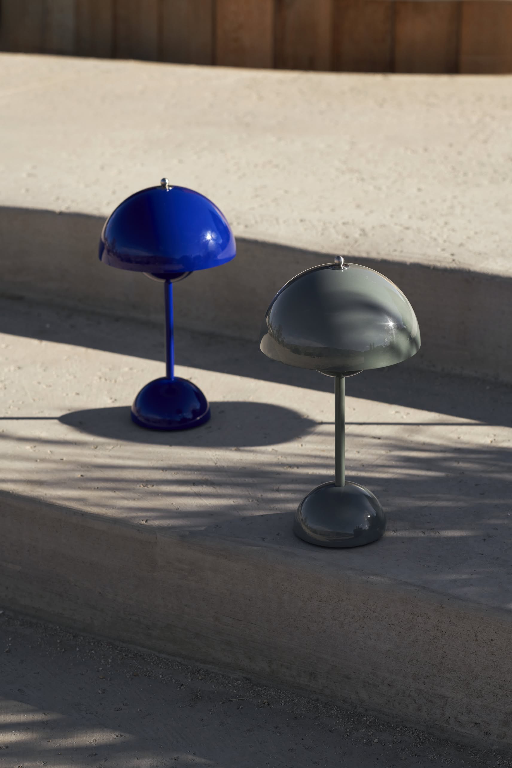 Two &Tradition Flowerpot VP9 Portable Table Lamps, one in glossy blue and one in gray, rest on outdoor concrete steps, casting soft shadows in sunlight. The portable design lets you easily move them for any occasion.