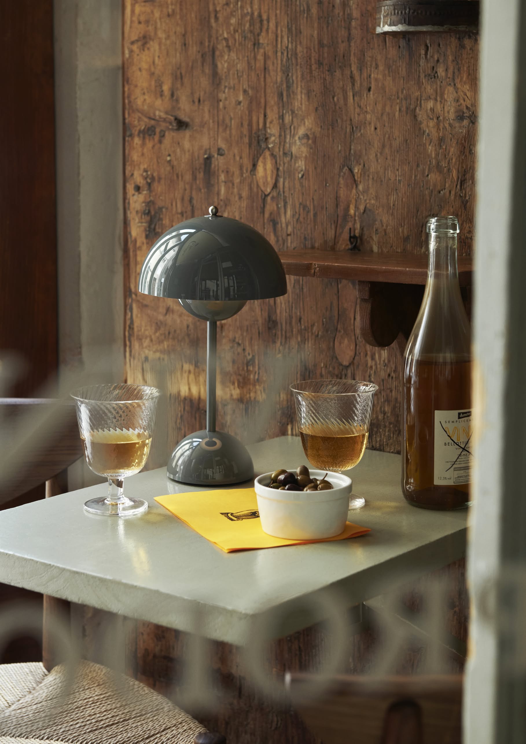 A small table with two glasses of wine, a bottle, a bowl of olives, an &Tradition Flowerpot VP9 Portable Table Lamp by &Tradition, and a yellow notebook sits in a rustic wooden interior.