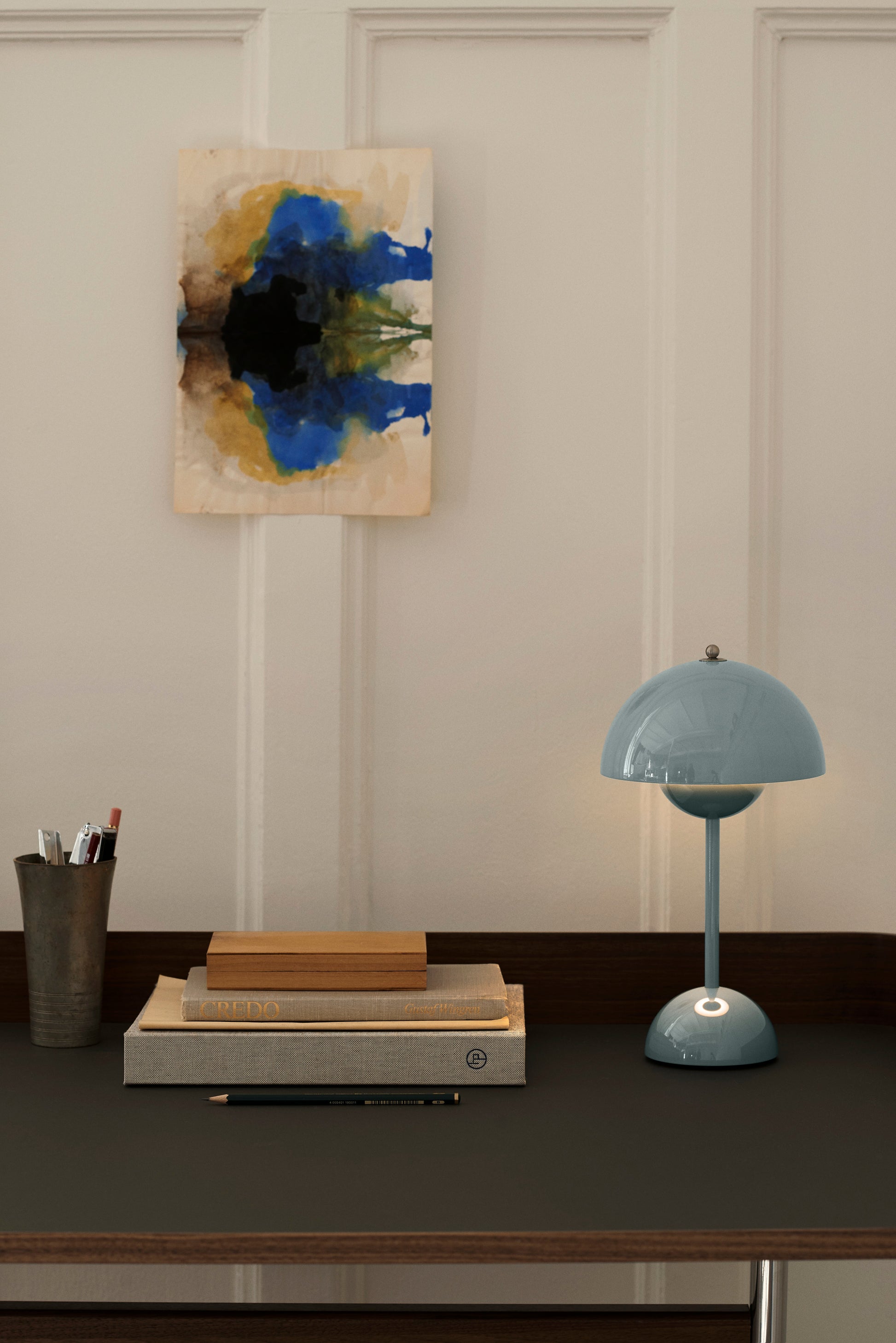 A minimalist desk with a pen cup, stacked books, a pencil, and the &Tradition Flowerpot VP9 Portable Table Lamp. An abstract black, blue, and brown ink artwork hangs on the white paneled wall above. A USB charging cable lies nearby.