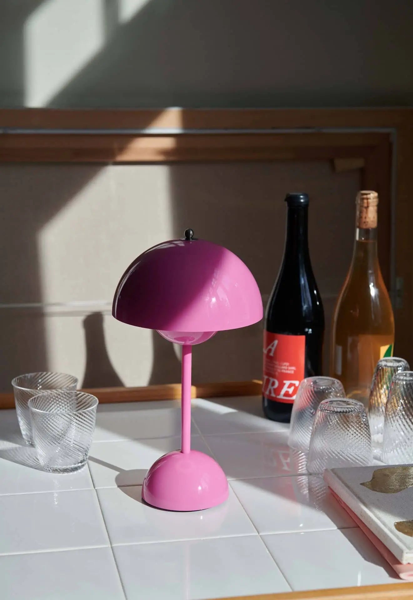 The &Tradition Flowerpot VP9 Portable Table Lamp in pink sits on white tiles amid textured glass cups, wine bottles, and a closed book. Sunlight and shadows accent the scene, while a USB charging cable highlights its portable design.