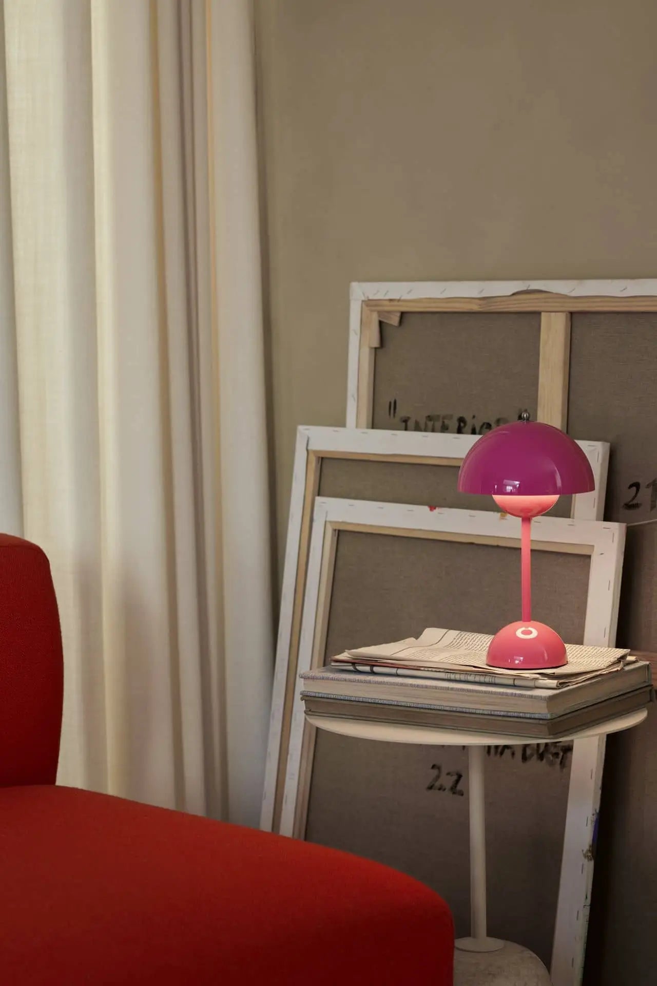 An &Tradition Flowerpot VP9 Portable Table Lamp by &Tradition, in modern pink, with a USB charging cable, rests atop stacked books next to a red chair. Large blank canvases lean against the wall in a softly lit room with white curtains.