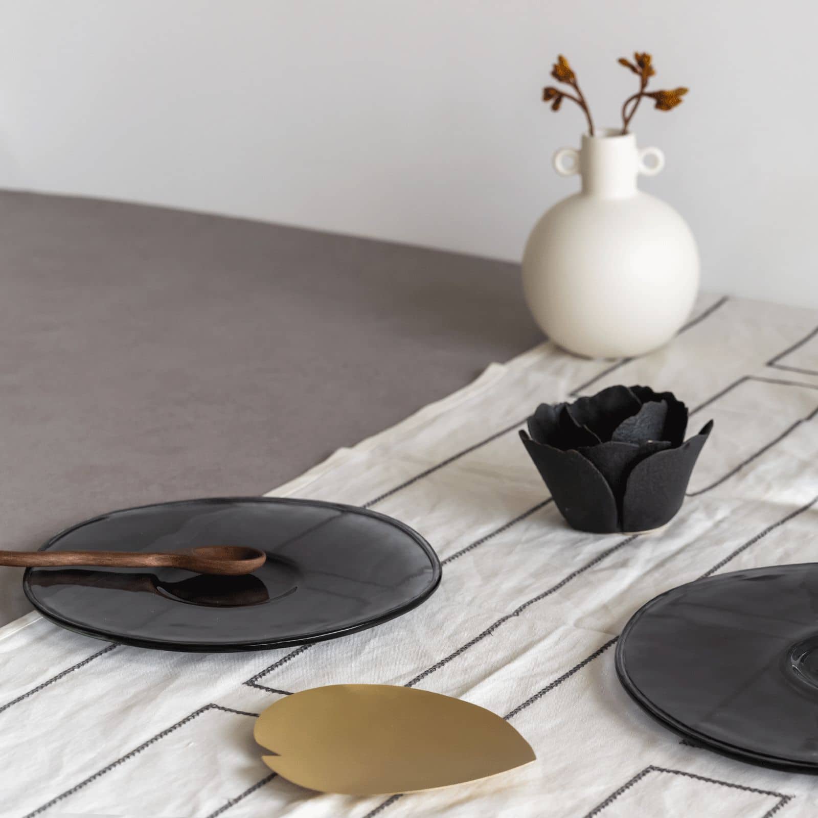 A minimalist table setting features the Urban Nature Culture Breakfast Plate Recycled Glass, a wooden spoon, gold leaf decor, and a black flower bowl; a white vase with dried flowers sits on a striped runner—ideal for sustainable living.