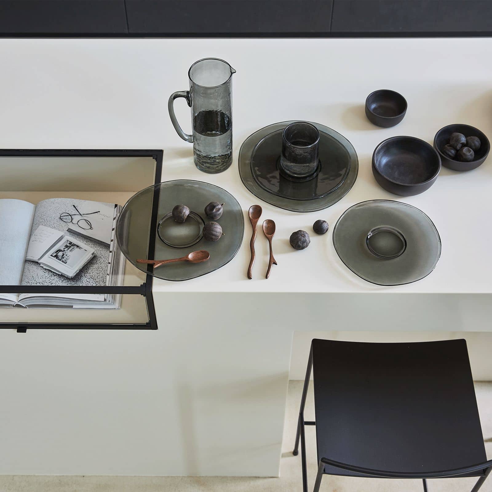 A minimalist kitchen counter features Urban Nature Culture Dinner Plates, recycled glass bowls, a water pitcher, wooden spoons, and black plums. An open book and a photo sit beside a black stool, adding a subtle rustic touch.
