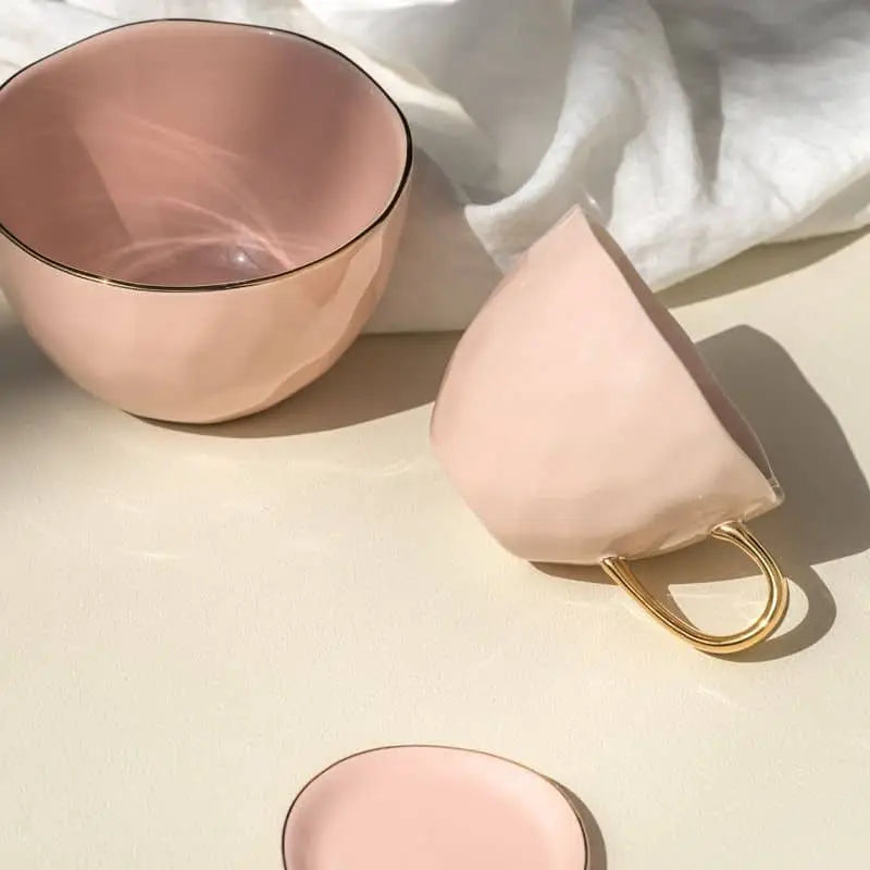 A pale pink Urban Nature Culture Good Morning Bowl glows in soft sunlight, its gold handle shining beside a matching saucer—perfect for your morning meal with gentle shadows and white fabric behind.