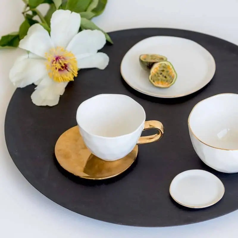 A black round tray holds a white teacup with a gold handle and saucer, the Urban Nature Culture Good Morning Bowl in soft pink pastel, a small plate, a dessert plate with passion fruit slices, and a white flower with green leaves.
