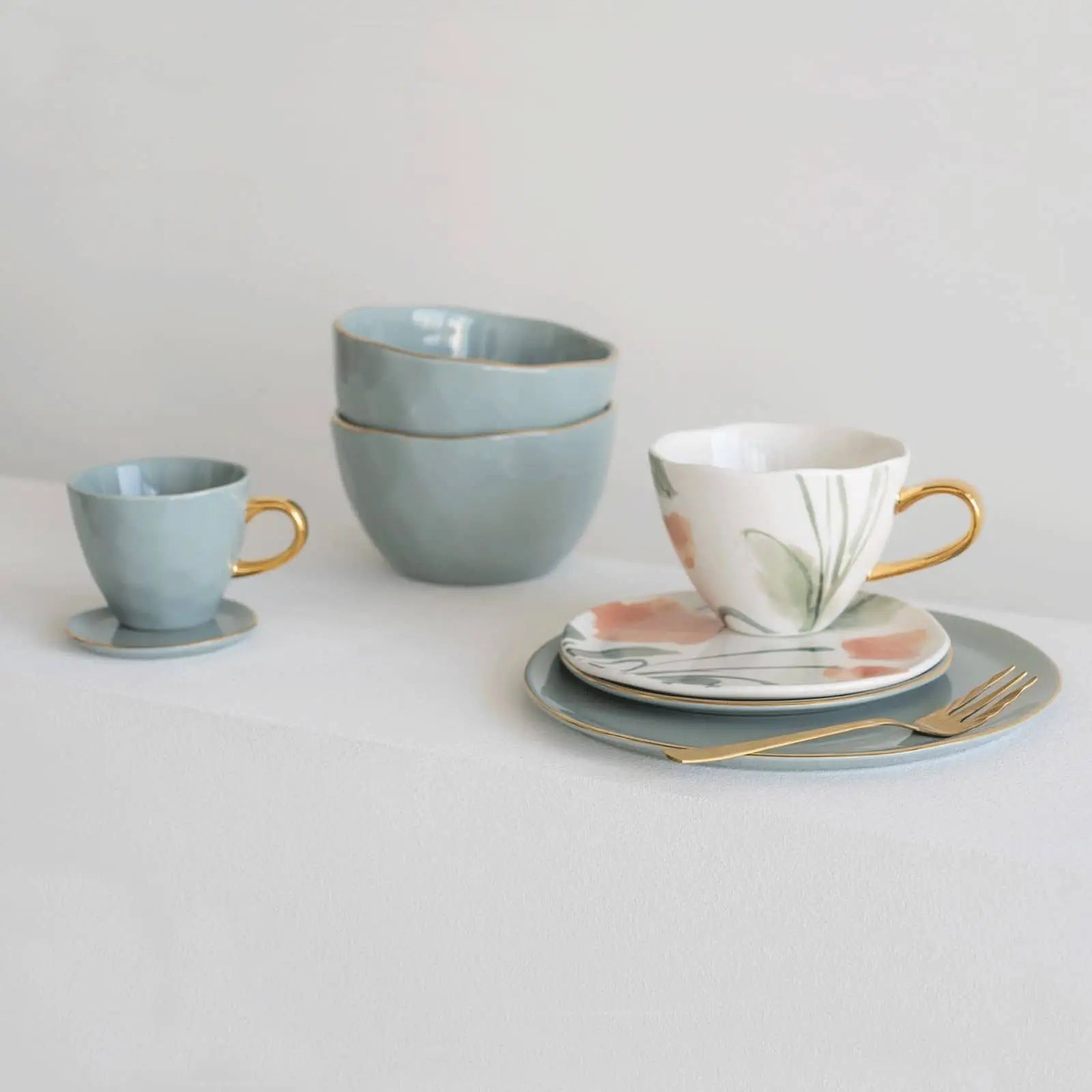 A minimalist table setting with blue ceramic bowls and cups, a floral-patterned teacup with gold handle, saucer, gold fork, and the Urban Nature Culture Good Morning Breakfast Plate—all resting on a white tablecloth in soft pastels.
