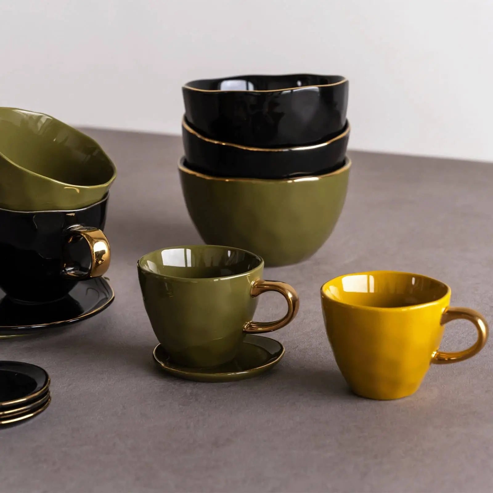 The Urban Nature Culture Good Morning Cup Mini by Urban Nature Culture is a ceramic cup in olive green, black, and mustard yellow, displayed on a gray surface with a light background alongside matching saucers and bowls.