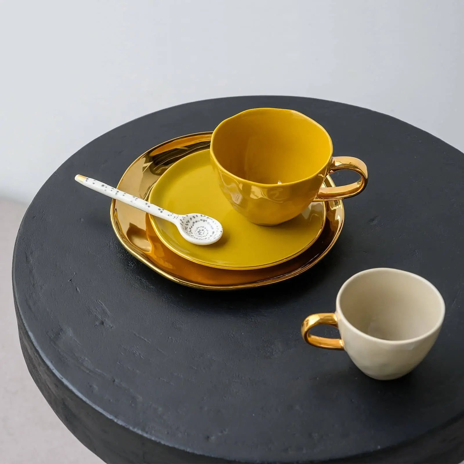 Two Urban Nature Culture Good Morning Cup Mini ceramic cups, one yellow and one cream, with gold handles sit on a black table. The yellow cup is on a matching saucer with a gold rim and comes with a decorative white spoon.