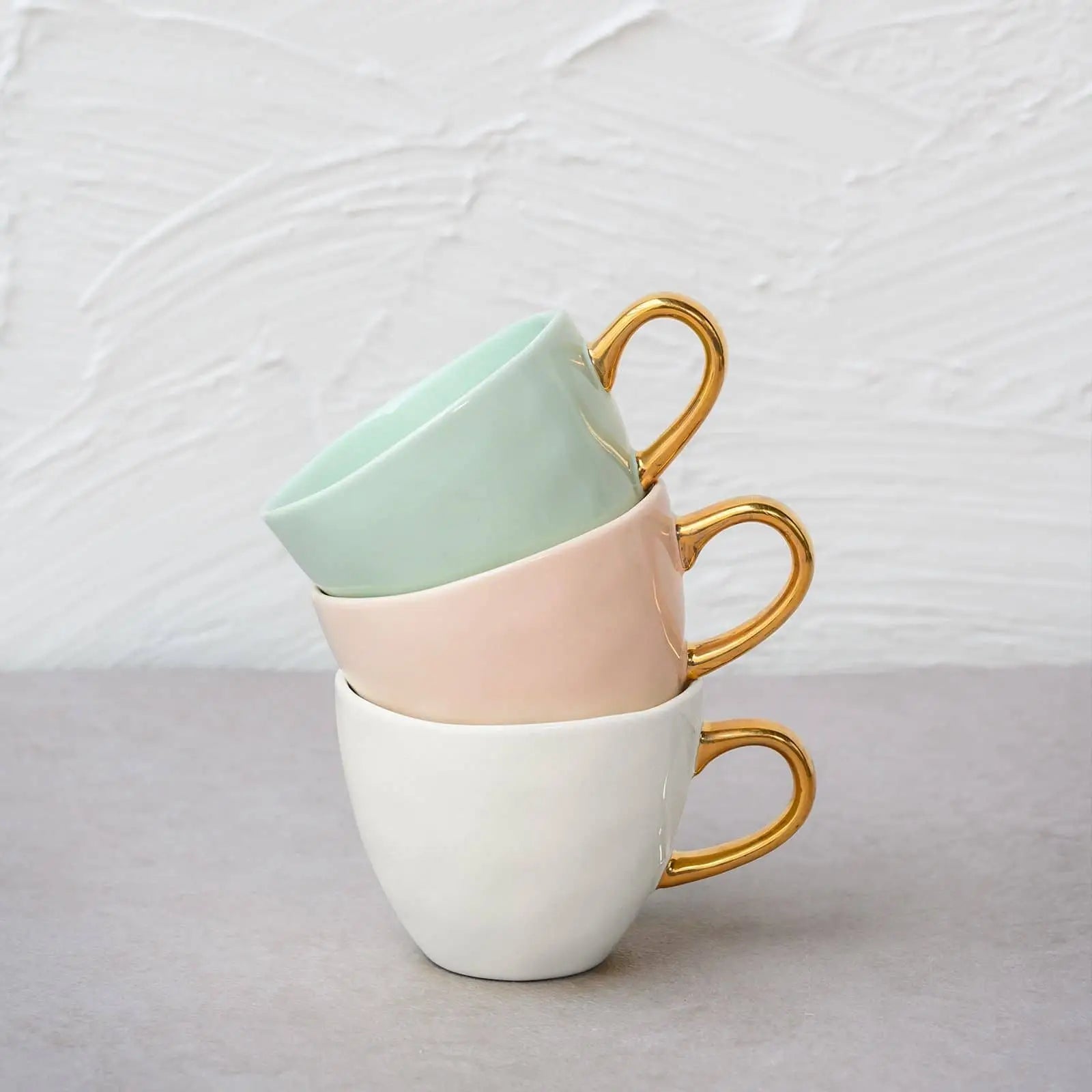 Three Urban Nature Culture Good Morning Cup Mini ceramic cups by Urban Nature Culture—in mint green, blush pink, and white with gold handles—are stacked on a gray surface with a textured white background.