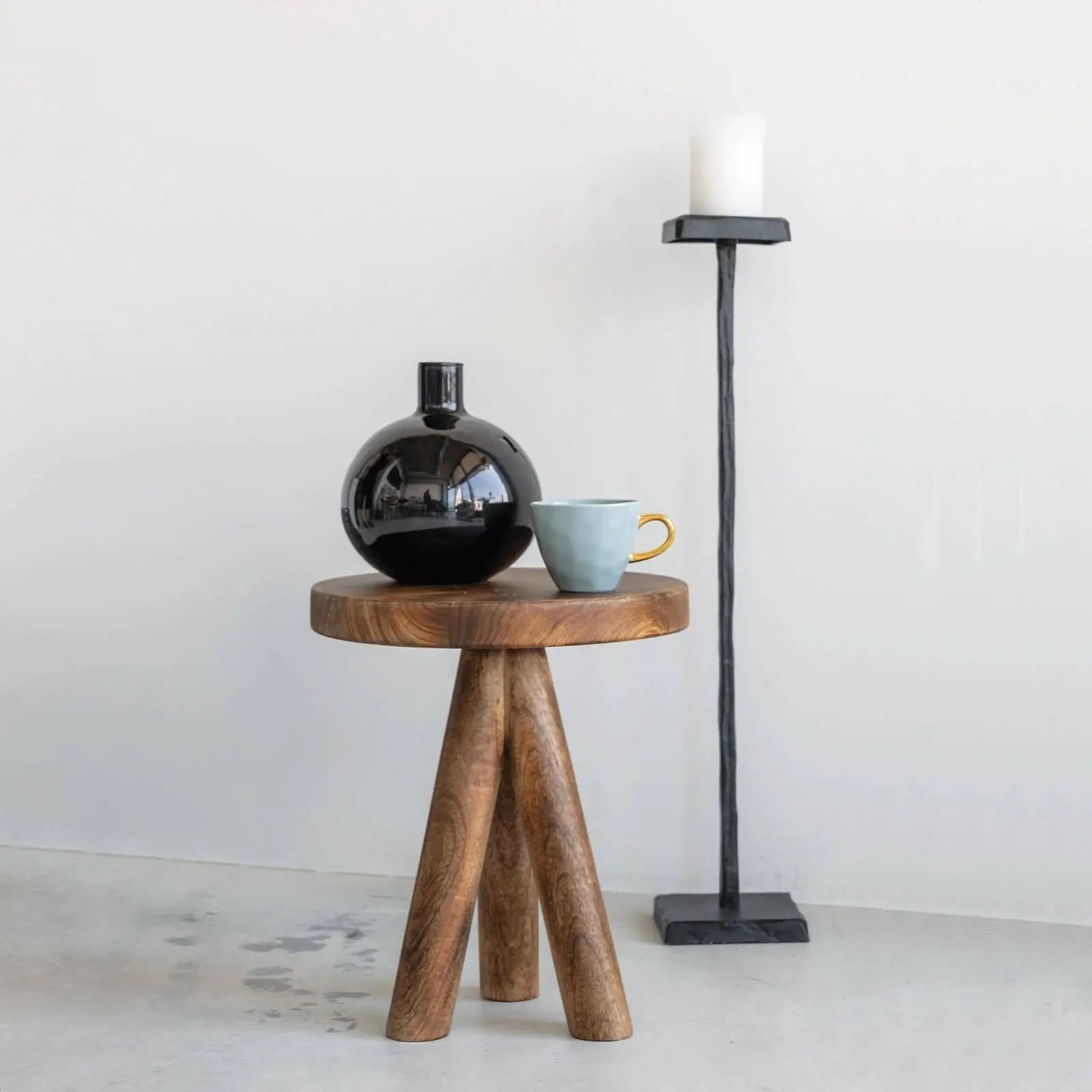 A small wooden tripod table holds a black round vase and an Urban Nature Culture Good Morning Cup Mini. Beside it stands a tall black metal candle holder with a white pillar candle, set against a plain white wall.