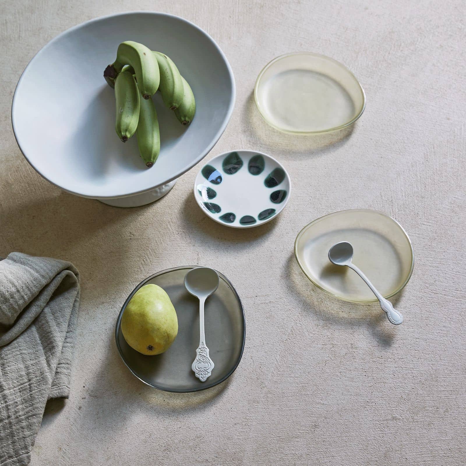A minimal table setting featuring bananas in a white bowl, a pear on an irregular plate, two Urban Nature Culture Plate Recycled Glass Frost, a small patterned dish, and a folded grey napkin on a light surface with earthy tones.