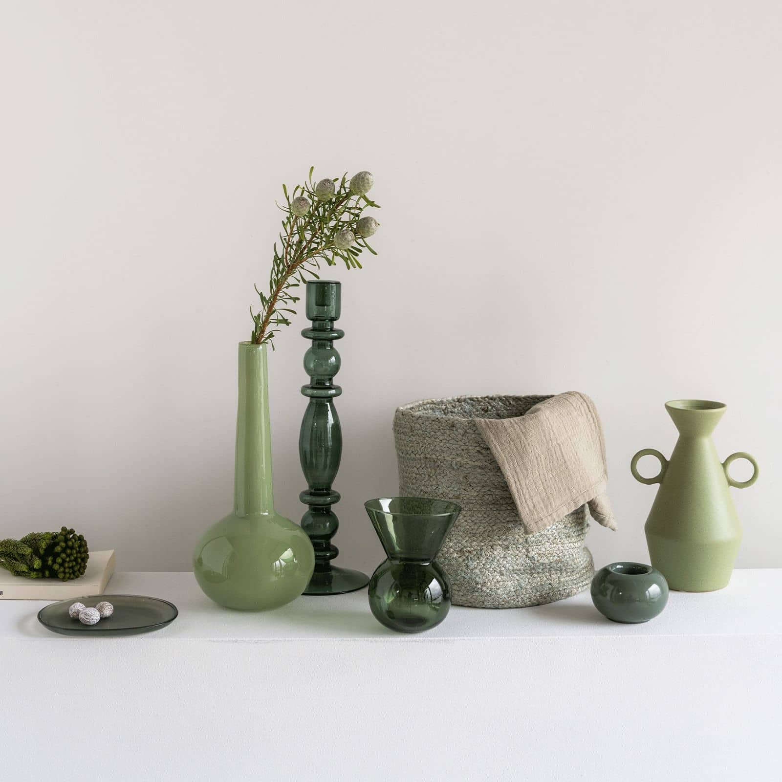 A selection of decorative green vases, bowls, and vessels in earthy tones sits on a white surface. The display includes the Urban Nature Culture Plate Recycled Glass Frost, an irregular plate by Urban Nature Culture, paired with ceramics and glassware.