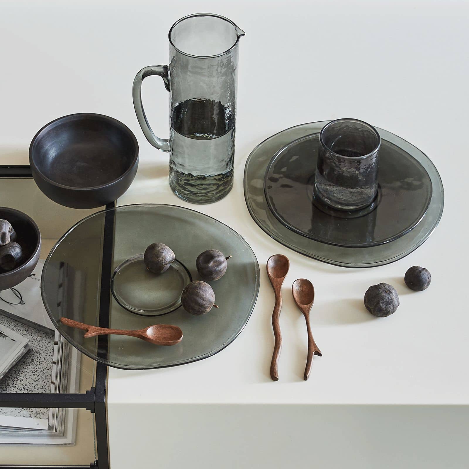 A minimalist table setting features smoky gray plates, a handmade pitcher, dark ceramic bowl, wooden spoons, Urban Nature Culture Tumbler Recycled Glass, and small round fruits—all arranged on white for an eco-friendly design.