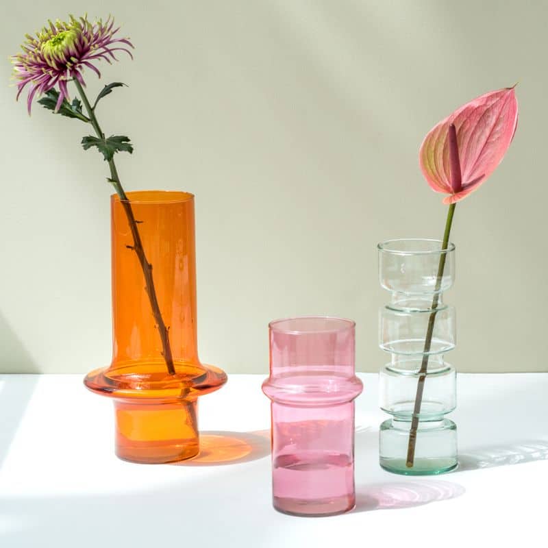 Three Urban Nature Culture vases—Paprika orange, pink, and clear—rest on a white surface. The orange recycled glass vase holds a purple flower, the clear one holds a pink flower, and sunlight casts soft shadows.