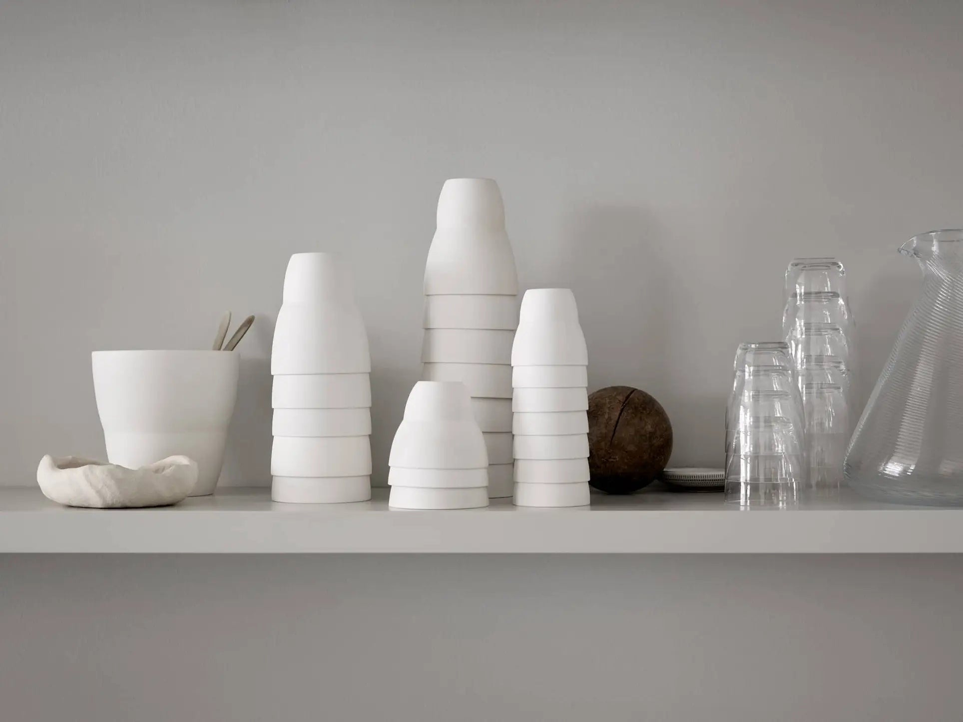 A minimalist shelf showcases Vipp Vipp201 Espresso Cups, Set of 2 by Vipp, along with porcelain containers, glass tumblers upside down, a glass pitcher, and a round brown object against a light gray wall.