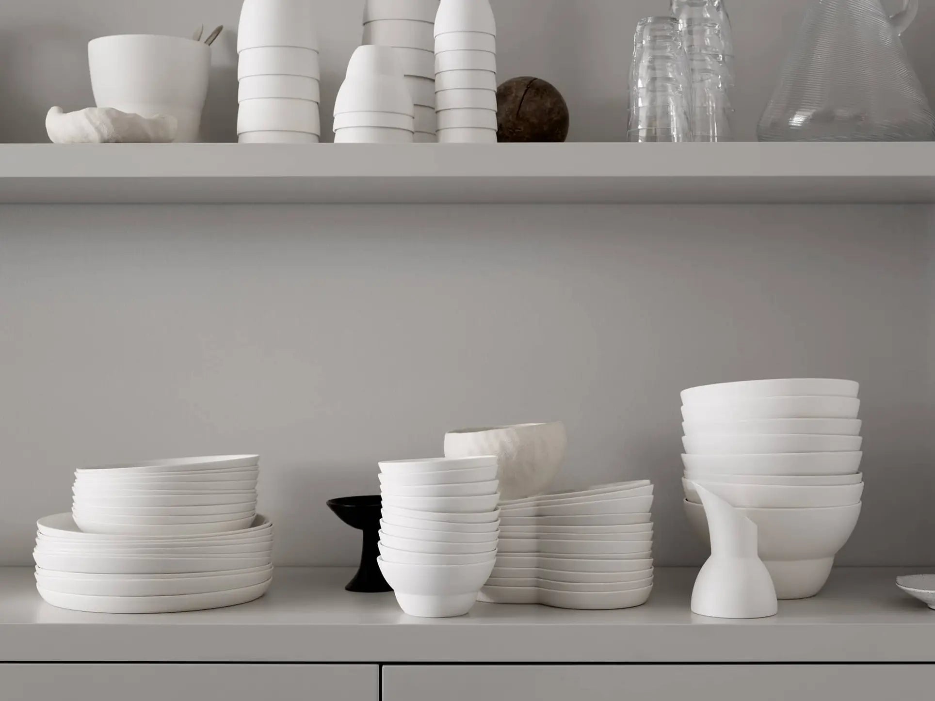 White ceramic plates, bowls, and a Vipp Vipp201 Espresso Cups set sit neatly stacked on a shelf against a light gray wall, with glassware and Danish porcelain pieces completing the minimal, organized arrangement above.