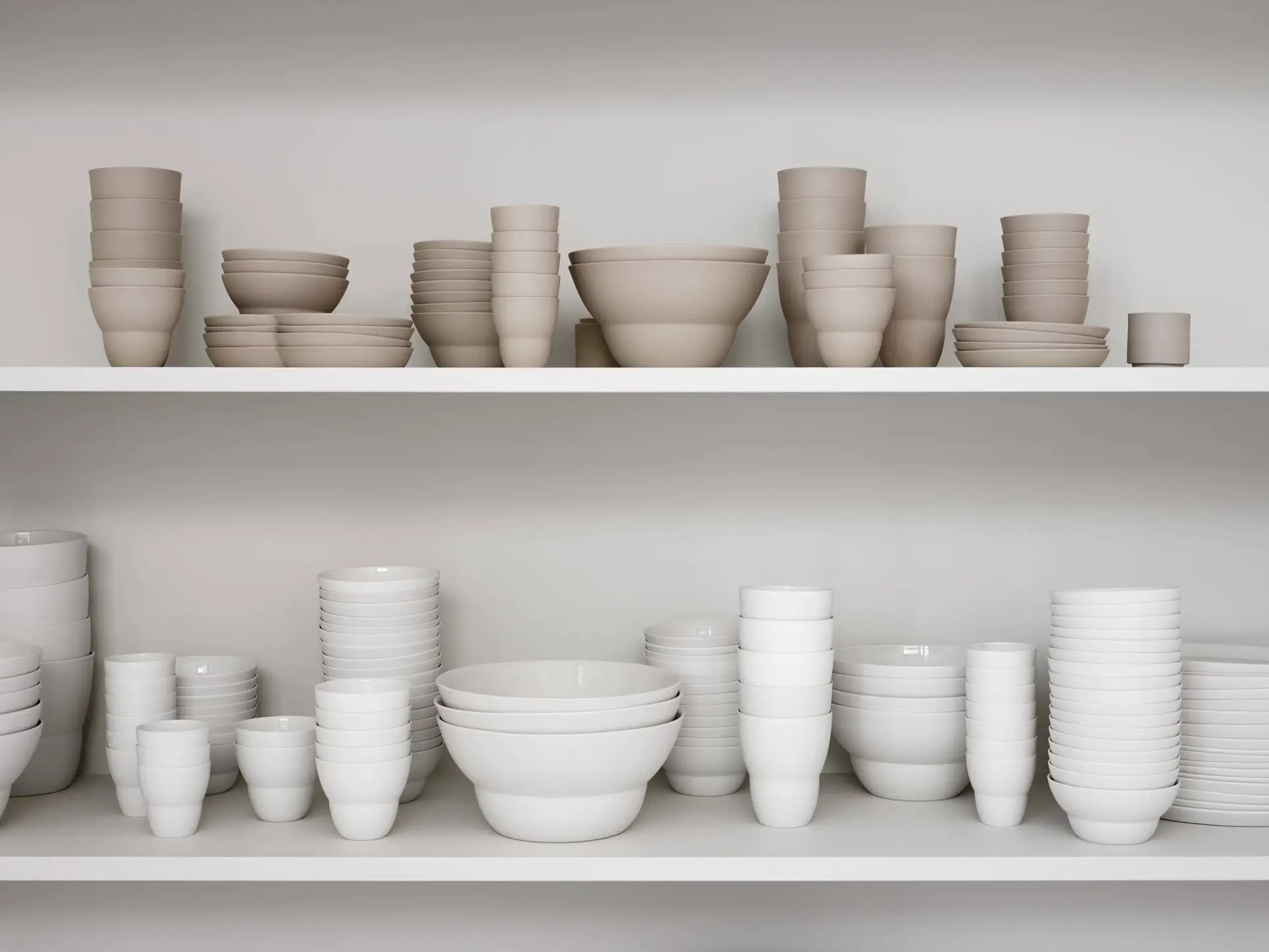 Two white shelves display neatly stacked Vipp Vipp201 Espresso Cups, Set of 2 by Vipp in beige and white porcelain, alongside matching ceramic bowls and plates arranged in organized rows.