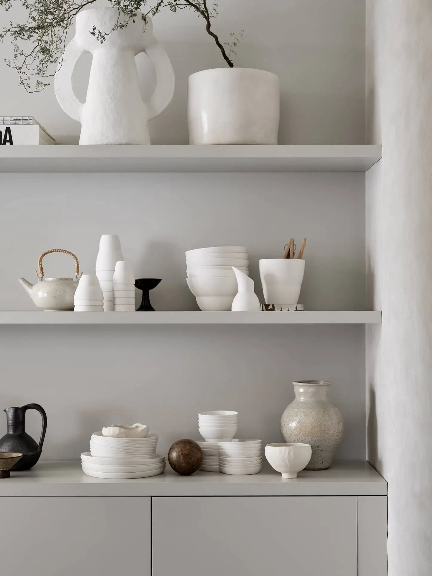 Minimalist kitchen shelves display white ceramics, Vipp Vipp201 Espresso Cups by Vipp, bowls, vases, and Danish pottery. A black pitcher, teapot, and brown sphere complete the calm atmosphere with neutral tones and modern decor.