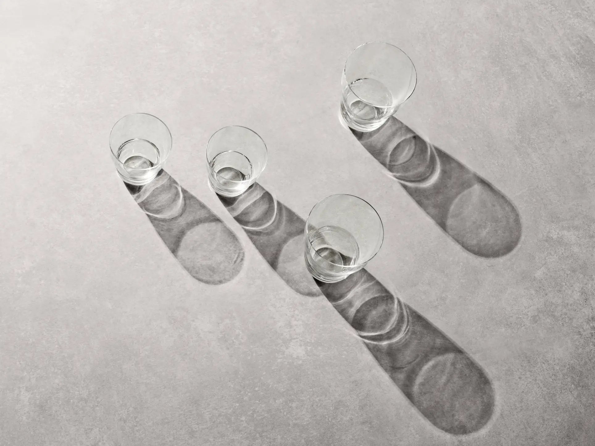 Two sets of Vipp Vipp240 5oz glasses by Vipp, crafted with mouth-blown glass, are arranged diagonally on a light gray surface, each casting long shadows to the right under bright, angled light.