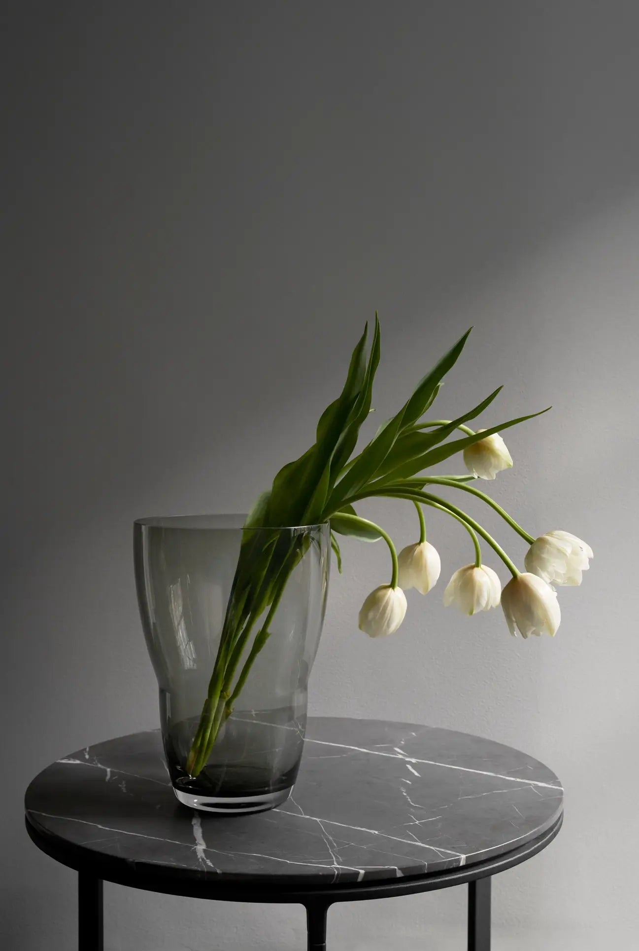 The Vipp Vipp248 Vase from Vipp, featuring smoke-colored glass and filled with white tulips and green leaves, is displayed on a round black marble table against a plain gray wall for a minimalist, elegant look.
