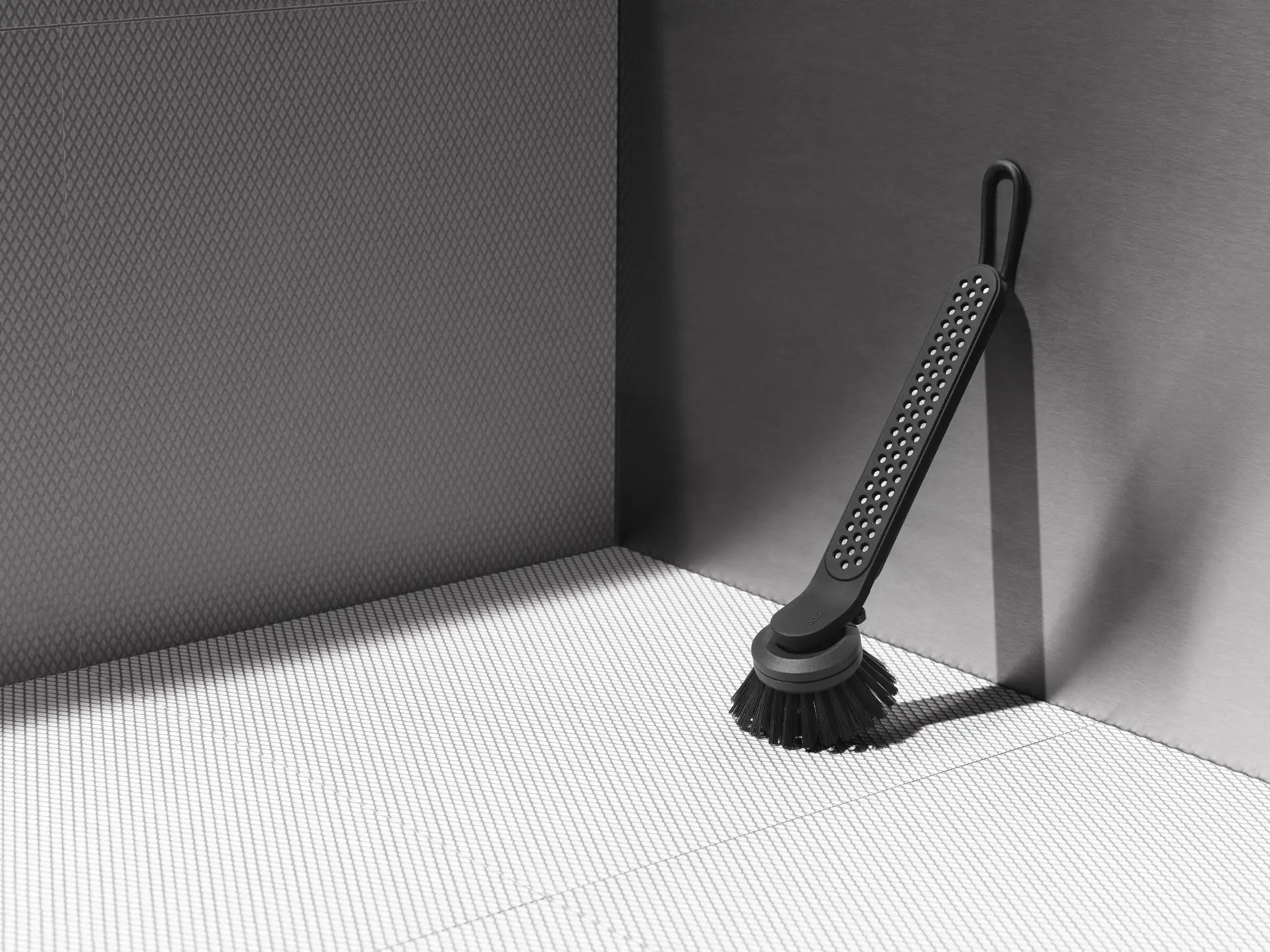 The Vipp Vipp280 Dishwashing Brush by Vipp, featuring a black long handle and replaceable brush head, stands upright on a tiled gray surface in a modern bathroom or kitchen.