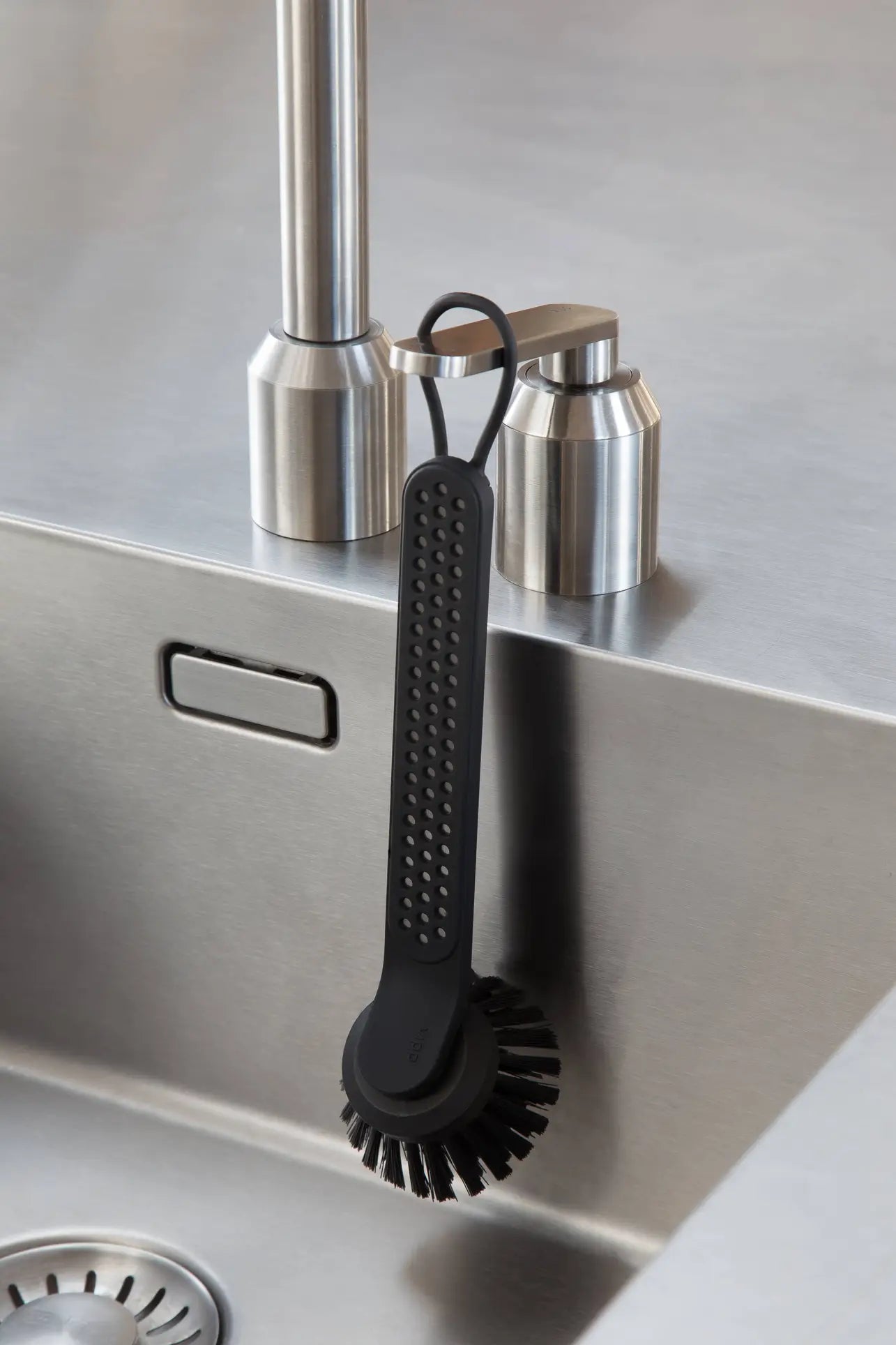 The Vipp Vipp280 Dishwashing Brush by Vipp, featuring a black textured handle and replaceable brush head, hangs from a soap dispenser on a stainless steel sink—enhancing any modern, efficient kitchen cleaning setup.