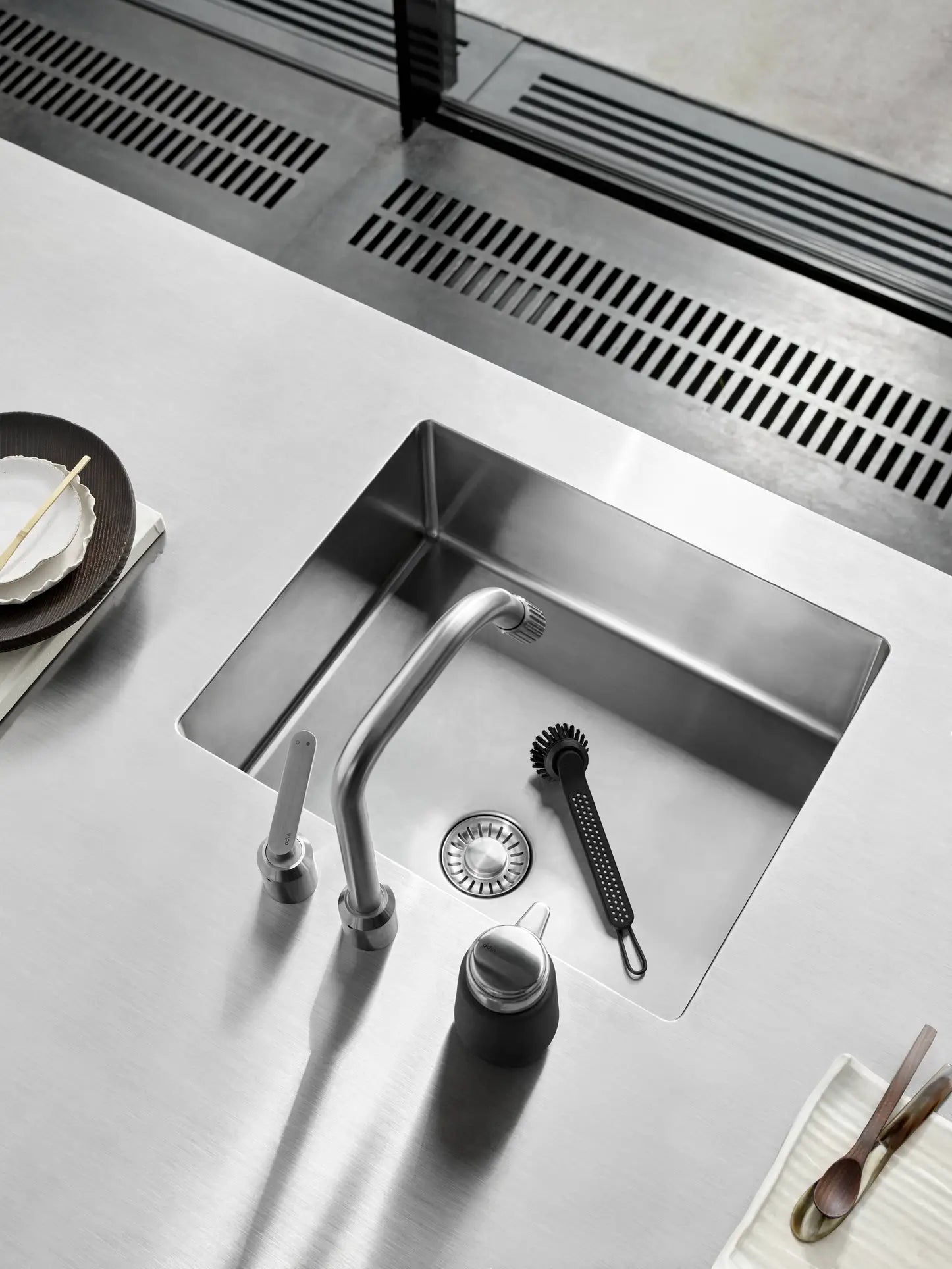 A modern stainless steel kitchen sink features a sleek faucet, a sink strainer, and the Vipp Vipp280 Dishwashing Brush by Vipp with a replaceable head. Plates, cutlery, and napkins are neatly arranged on either side for easy cleaning.
