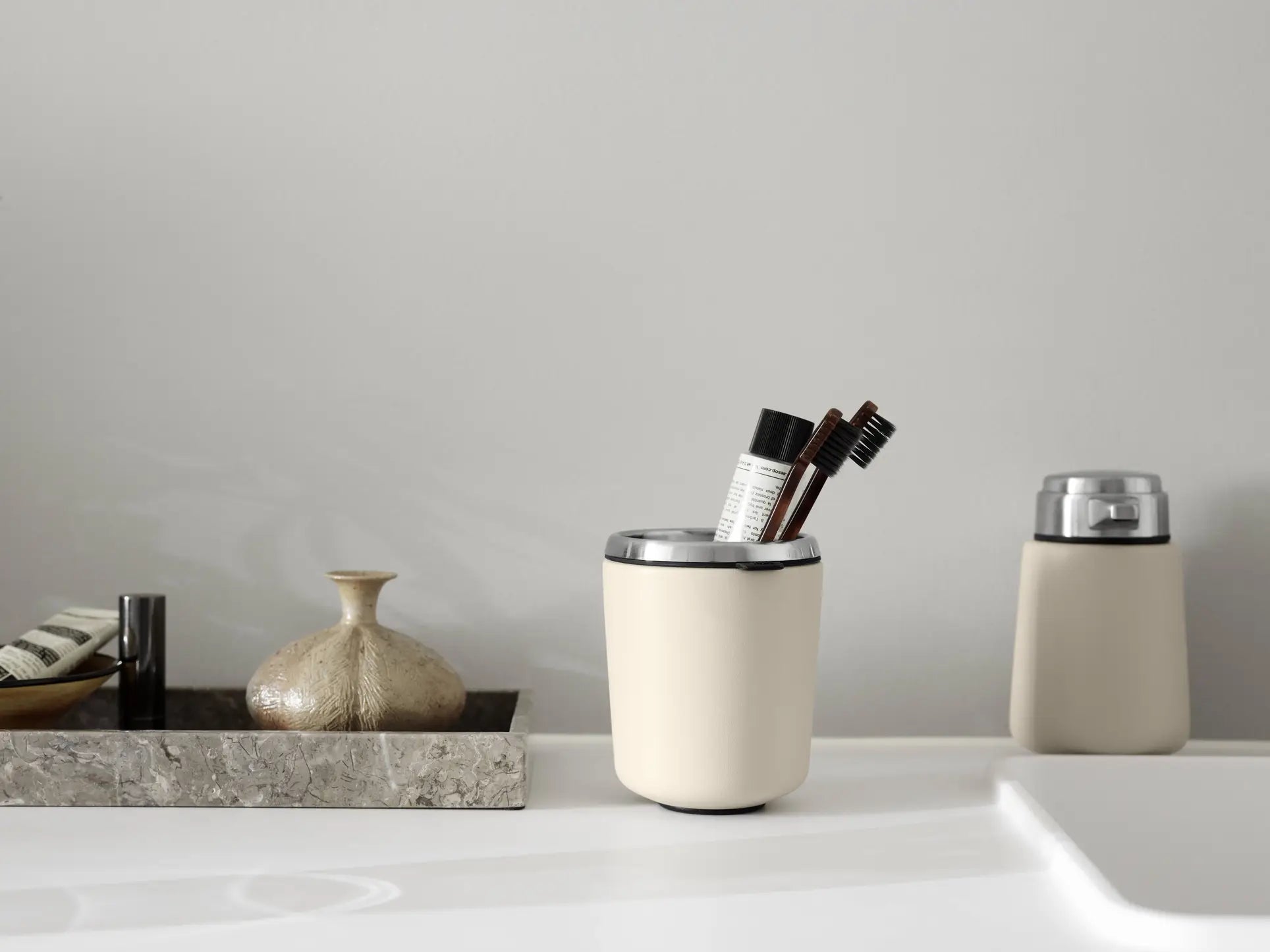 The Vipp Vipp7 Toothbrush Holder by Vipp, featuring a detachable rubber insert and filled with toothbrushes and a tube, stands on a white counter beside a stone tray with a vase and toiletries; a true bathroom essential. A soap dispenser is in the background.