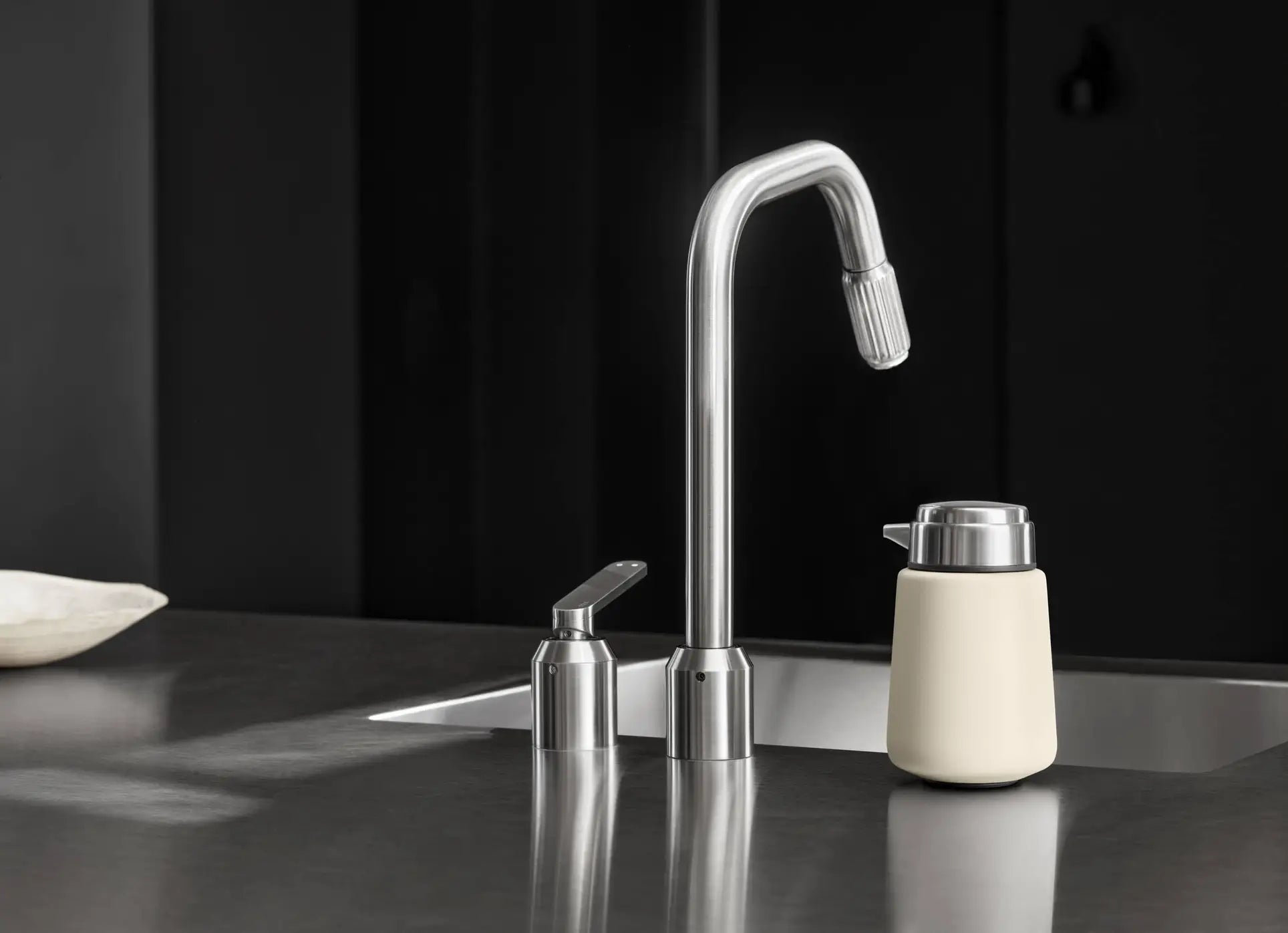 A modern stainless steel kitchen faucet with a matching Vipp Vipp9 Dispenser and beige container sit on a sleek, dark countertop in a minimalist kitchen, showcasing elegant accessory choices from Vipp.