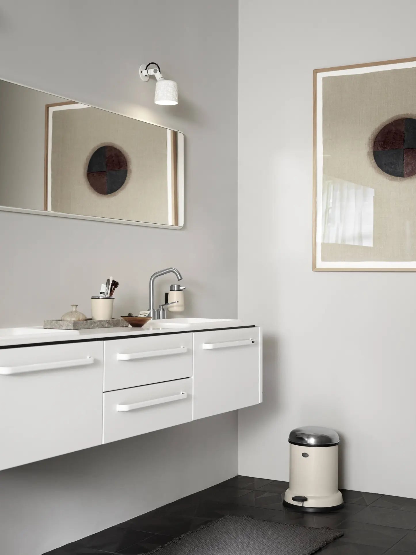 Modern bathroom with white floating vanity, minimalist sink, rectangular mirror, wall light, abstract art, small round trash bin, and black hexagonal floor tiles. The Vipp Vipp9 Dispenser by Vipp adds sleek style to the neutral color palette.