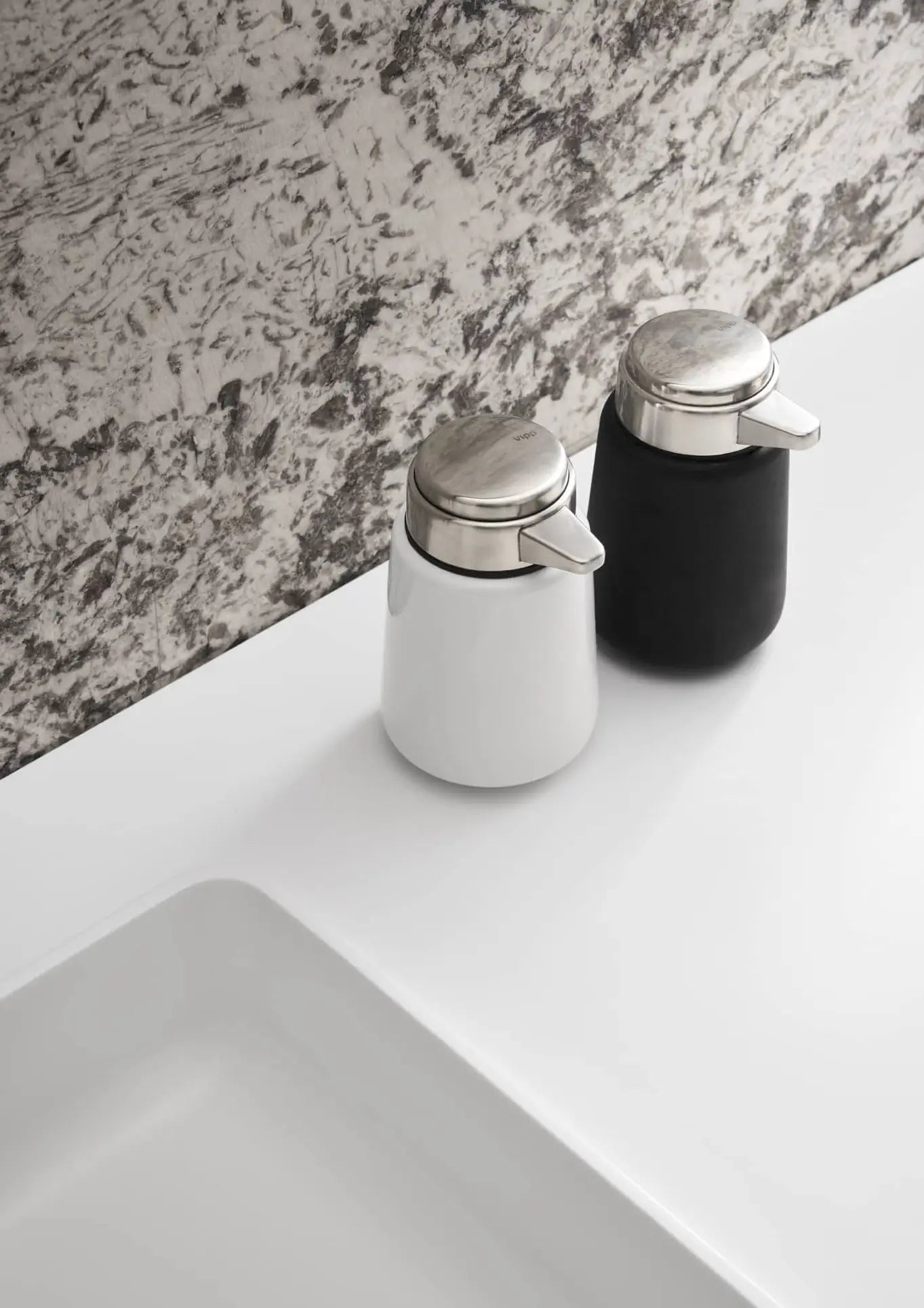 Two Vipp Vipp9 Dispensers by Vipp, one white and one black with silver tops, sit on a white bathroom counter by the sink against a gray-and-white patterned wall—ideal for adding sleek style to your kitchen or upgrading your bathroom.