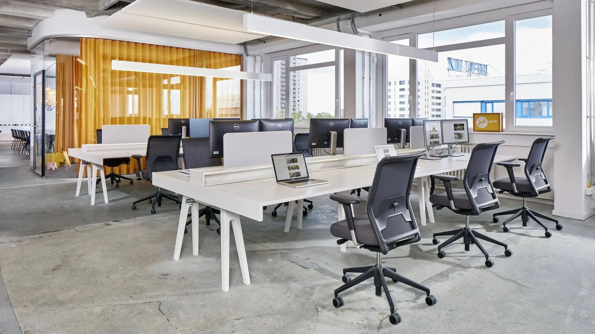 Modern open office with white desks, black chairs, monitors, and laptops. Large windows provide natural light. In the background, a yellow glass meeting room and a Vitra Eames Elephant, Small by Vitra add a charming decorative touch.