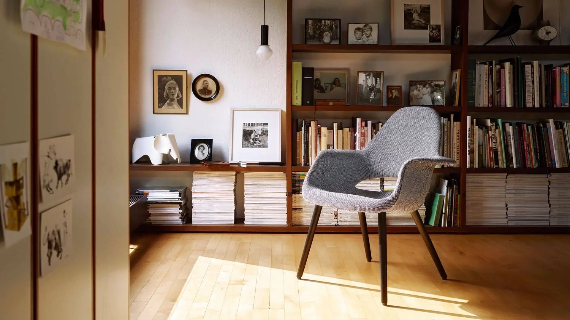 A modern gray chair sits in a sunlit room with wooden floors, bookshelves, framed photos and artwork, while a small Vitra Eames Elephant by Vitra adds a decorative touch next to stacked magazines.