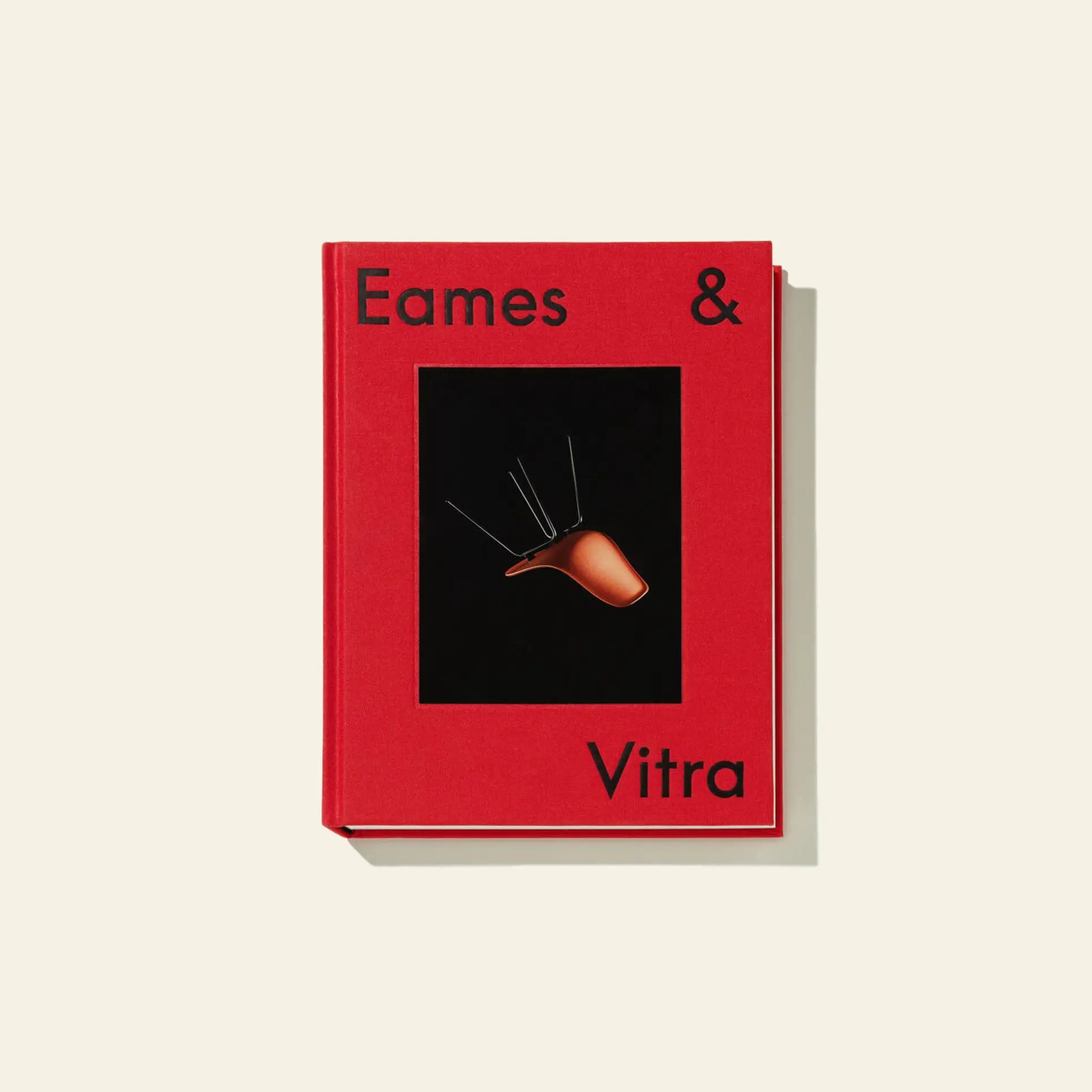 The Vitra Eames & Vitra Hardcover Book by Vitra has a red cover with black text and a brown Eames chair image on a black background, inspired by Charles and Ray Eames, casting a soft shadow on a light cream surface.