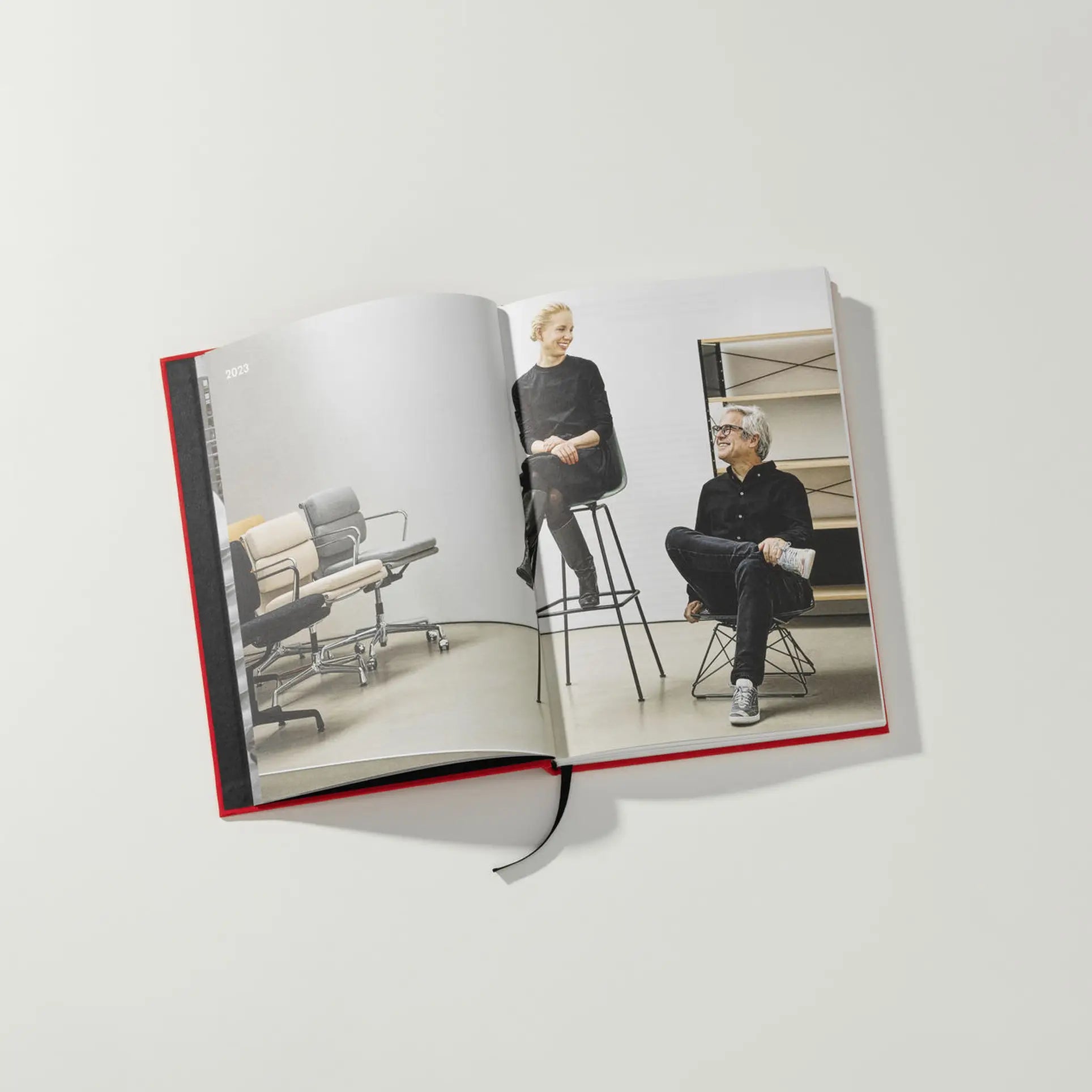 The Vitra Eames & Vitra Hardcover Book by Vitra features a photo of a woman on a tall stool and a man on a low chair, both dressed in black, surrounded by Eames furniture, with shelves and office chairs in the background. A ribbon bookmark is included.