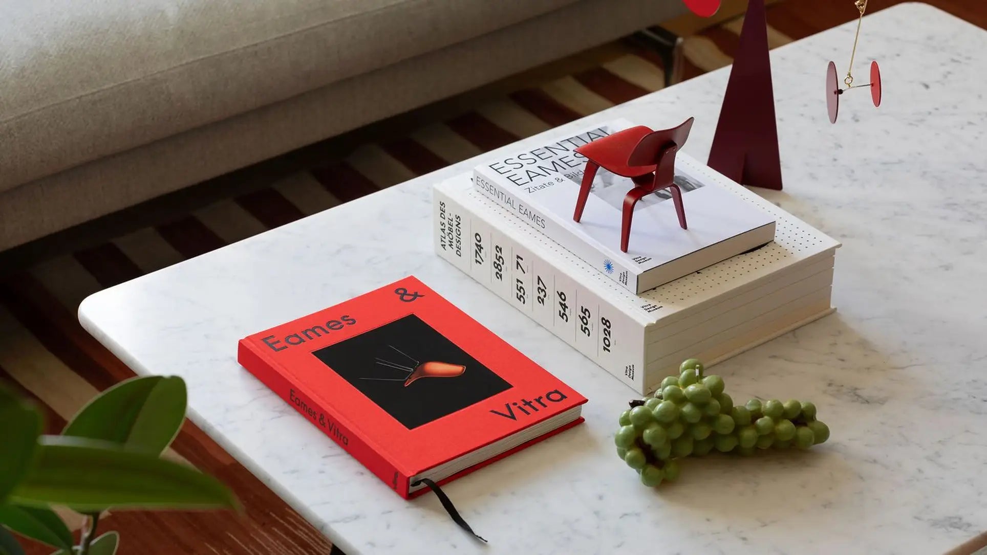 A white marble coffee table holds the Vitra Eames & Vitra Hardcover Book by Vitra, green grapes, stacked white books with Essential Eames on top, and a small red chair model—evoking the classic style of Charles and Ray Eames. Sofa and plants appear in view.