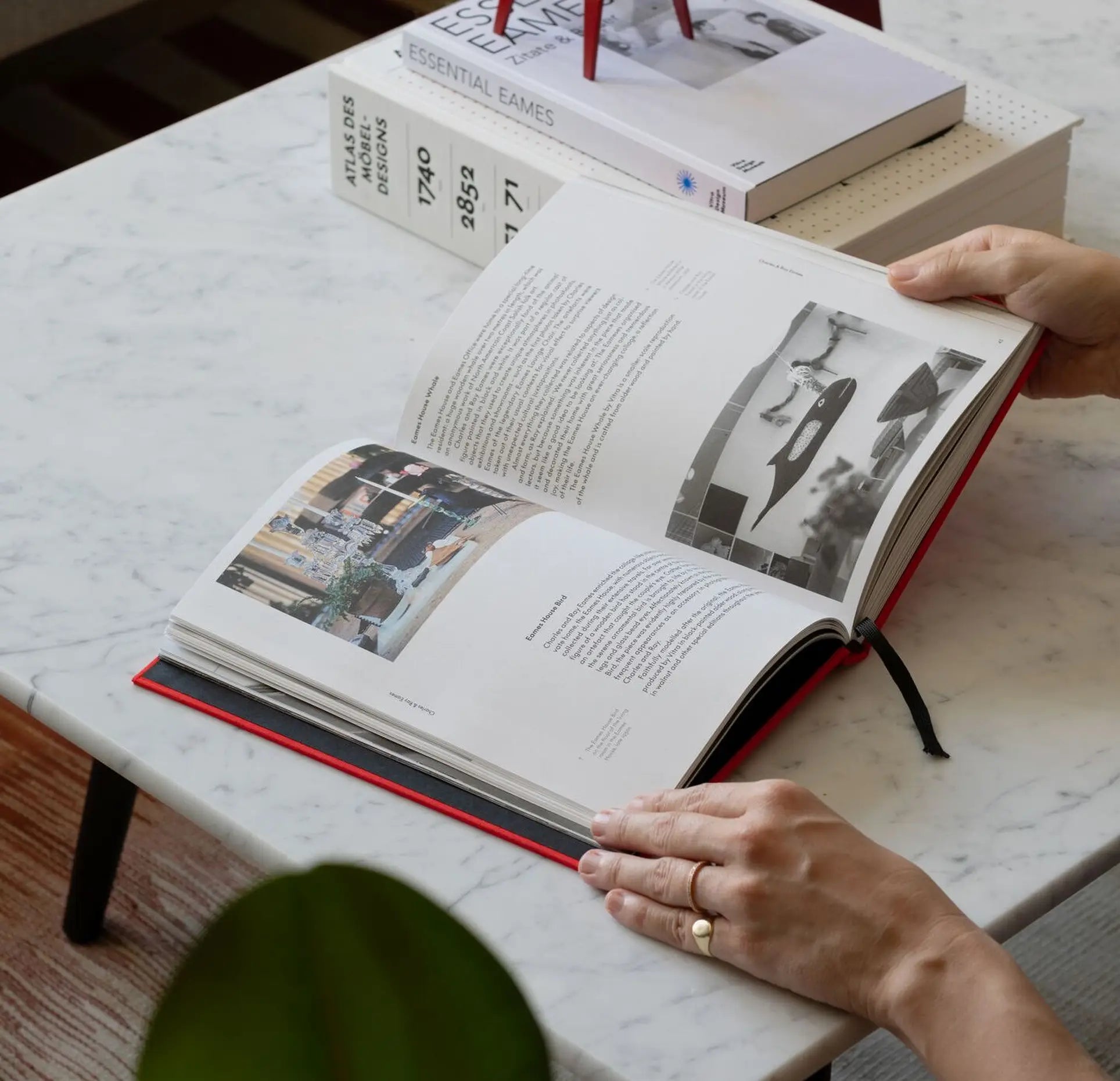 A person with a gold ring on their left hand reads an open book at a marble coffee table. Nearby, other books are stacked, including the Vitra Eames & Vitra Hardcover Book by Vitra about Charles and Ray Eames and their iconic furniture.
