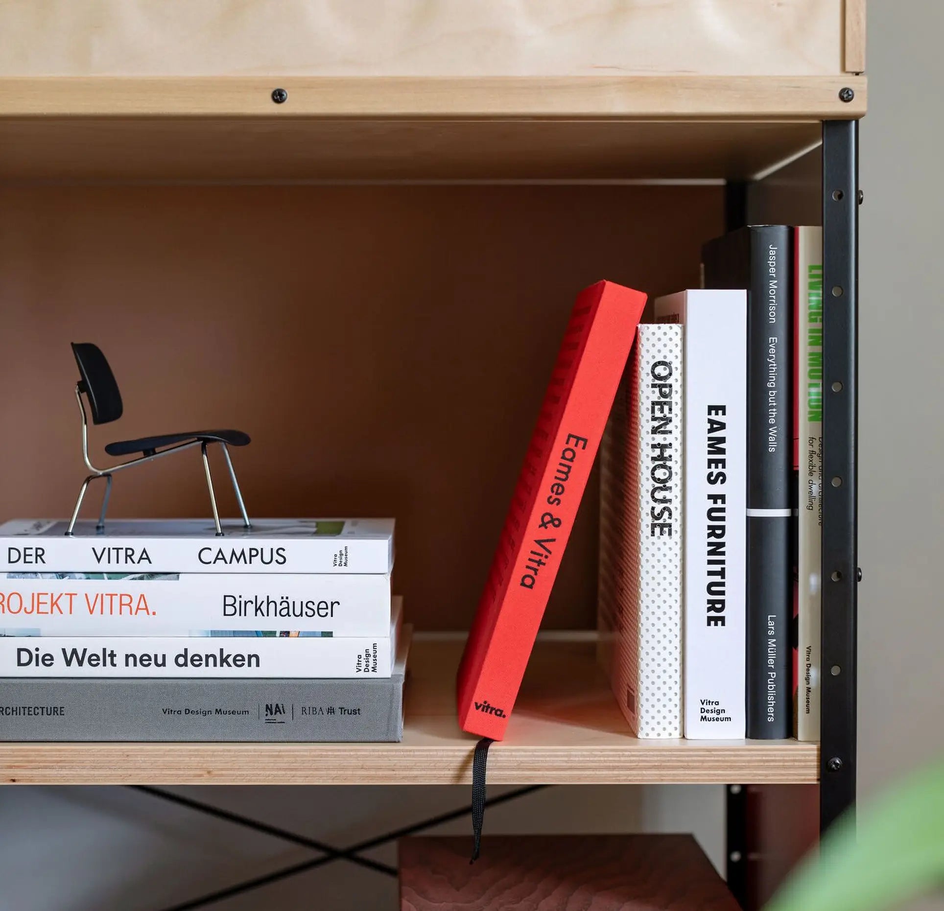 A close-up of a wooden bookshelf with neatly arranged books, including the Vitra Eames & Vitra Hardcover Book by Vitra. A small black Vitra Eames chair model sits atop a stack of books on the left shelf.