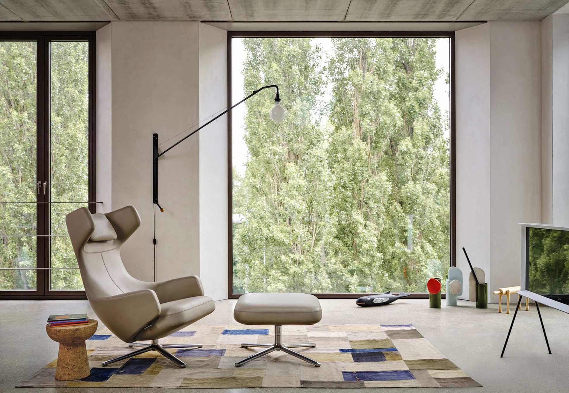 Modern living room with a beige lounge chair and ottoman, colorful rug, side table, Vitra Potence Wall Lamp - Deep Black, and large windows framing lush trees. Minimalist design and natural light create an inviting, contemporary space.