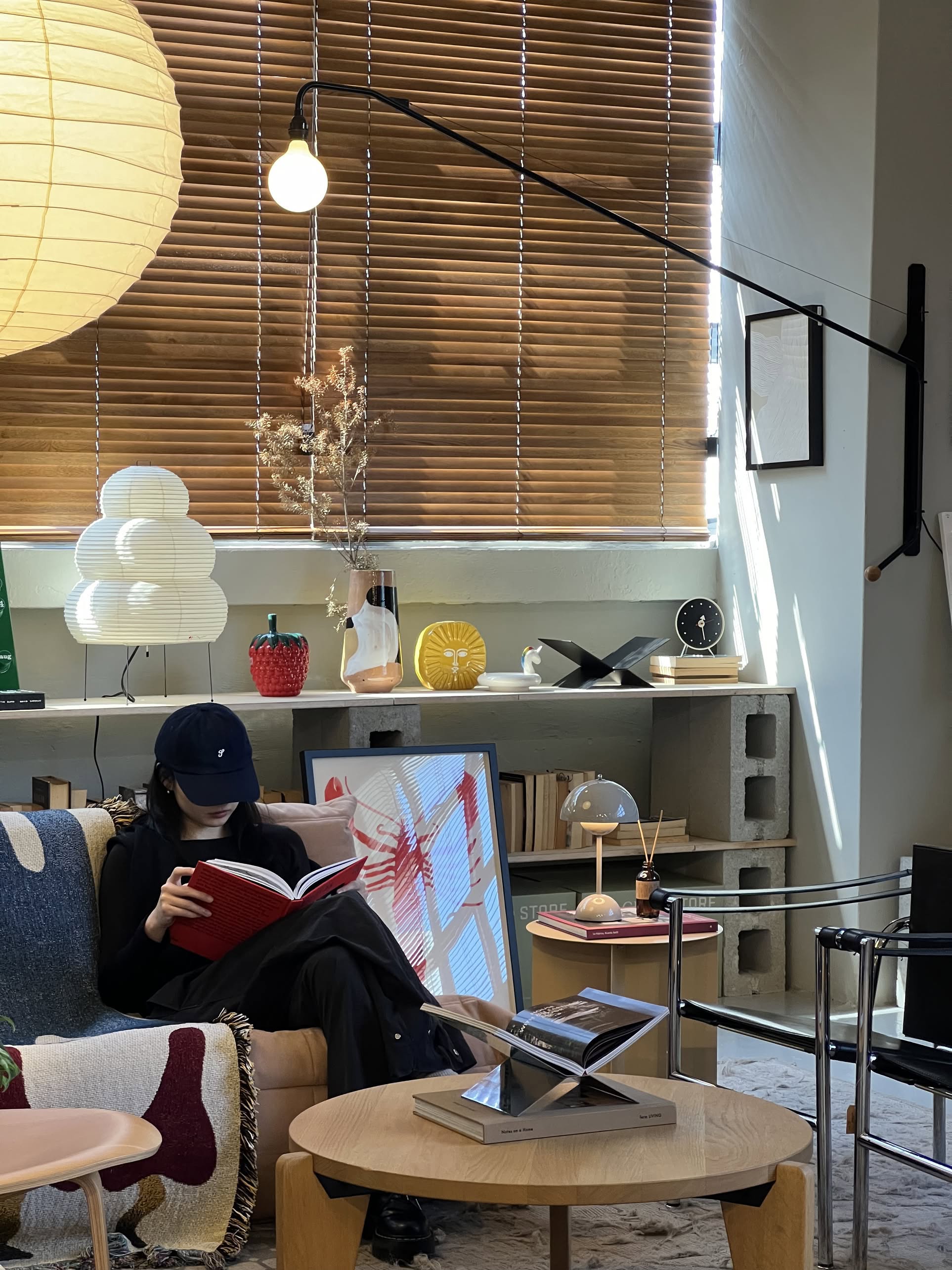 A person in dark attire and a cap reads a book on a couch in a modern living room with minimalist decor, wooden blinds, neat bookshelves, and the Vitra Potence Wall Lamp - Deep Black by Vitra enhancing the stylish ambiance.