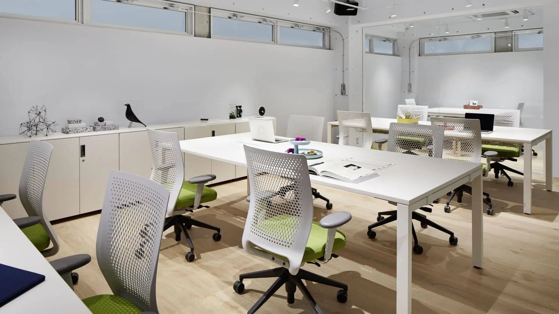 A modern, bright office with white walls, white desks, and green chairs. Large windows and light wooden floors complement stylish storage like the Vitra Rotary Tray by Vitra for organizing your belongings on desks and cabinets.