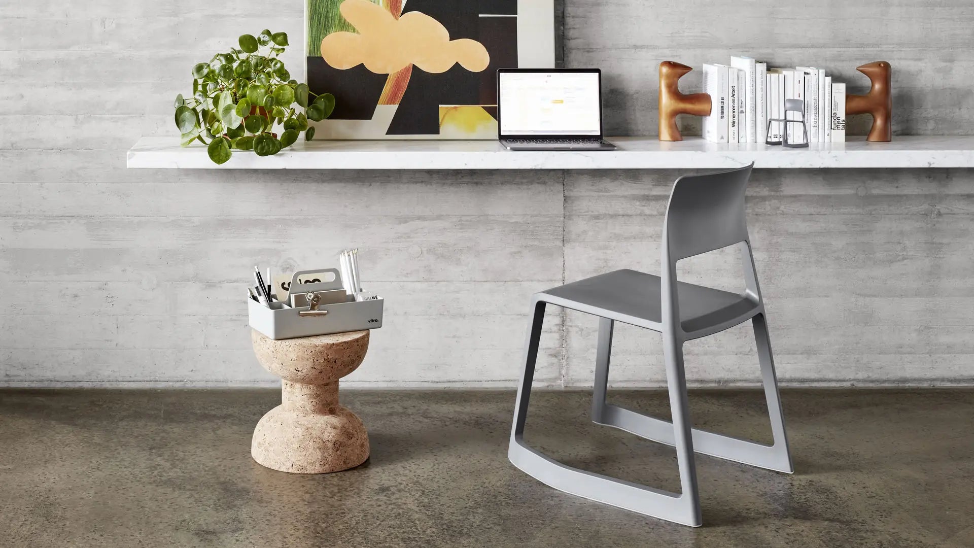 A modern workspace with a sleek gray Vitra chair, a cork stool holding office supplies in the Vitra Toolbox RE by Vitra—made from recycled ABS—plus a floating shelf desk with a laptop, books, plant, and abstract art against a concrete wall.