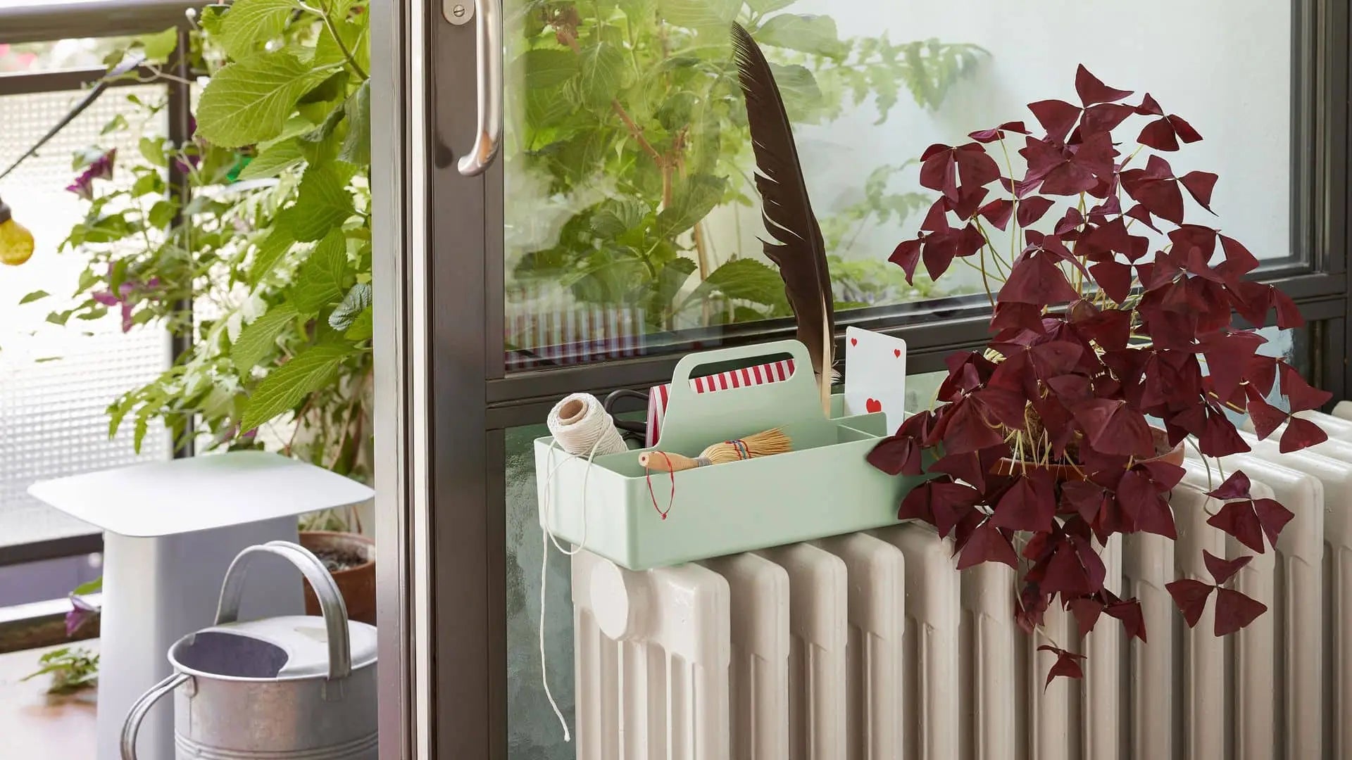 A mint-green Vitra Toolbox RE organizer by Vitra, crafted from recycled ABS plastic, rests on a white radiator beside a plant with purple leaves and a silver watering can, near a glass door overlooking lush green foliage.