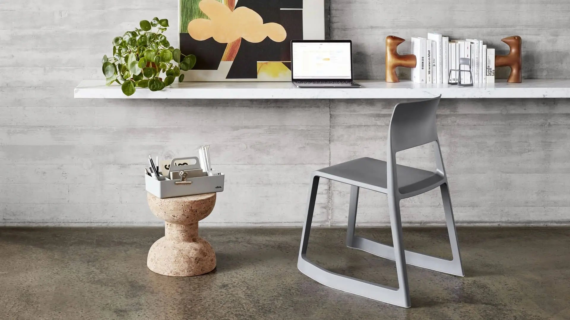 A minimalist workspace features a gray chair and floating desk with a laptop, books, and decor. On the concrete floor is the Vitra Toolbox RE by Vitra, made from recycled ABS plastic, next to a plant and cork stool with office supplies.