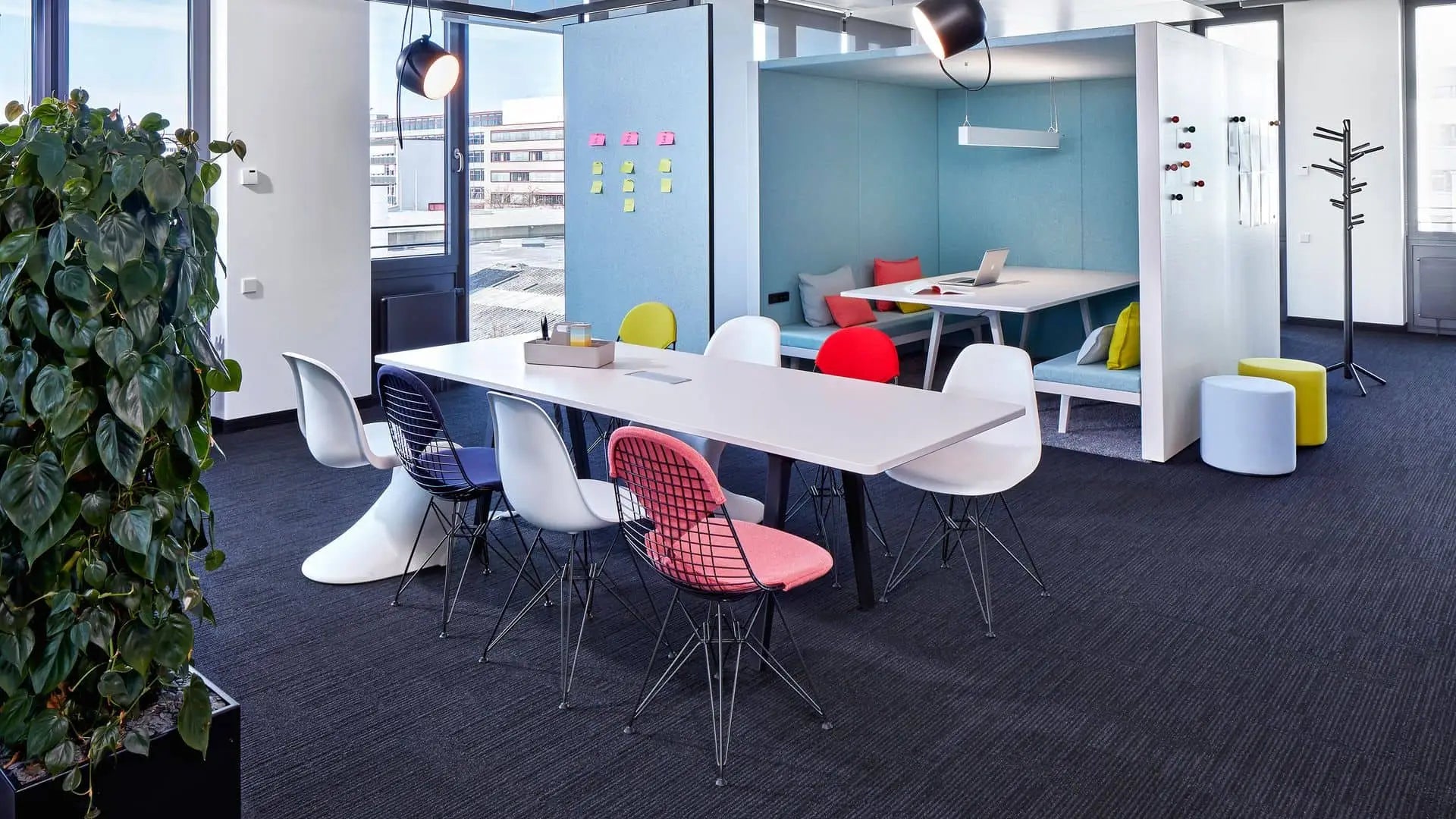 Modern office space with a long white Vitra Toolbox RE table by Vitra, colorful chairs, cubicle area with couches and workstations in the background, large windows, lush plants, and bright lighting.